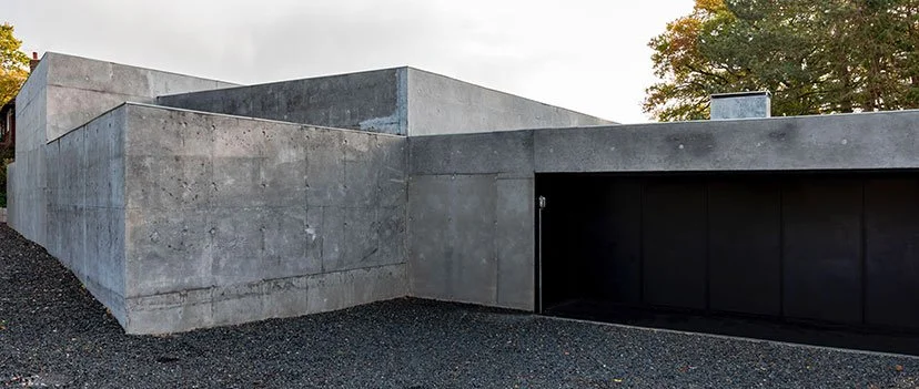 Modern concrete building with minimalist design, large black door, and gravel driveway, surrounded by trees.