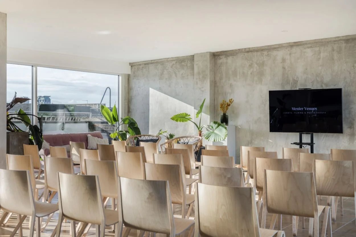 Empty event space with wooden chairs, large windows, potted plants, and a wall-mounted TV.