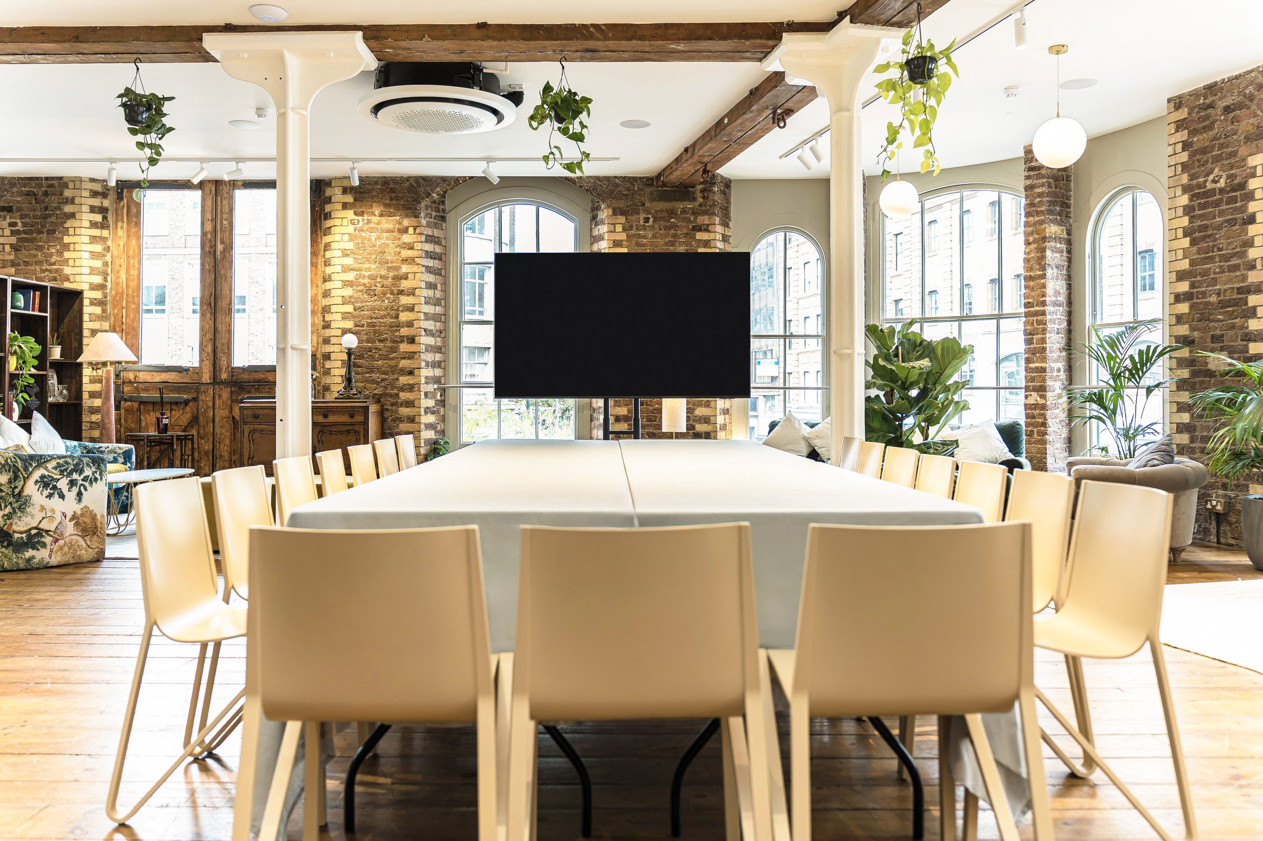 A large dining table with white chairs in an industrial-style room with exposed brick walls, large arched windows, wooden beams, and lots of natural light. The table has a white tablecloth, and a large black monitor is centered on it. There are vario