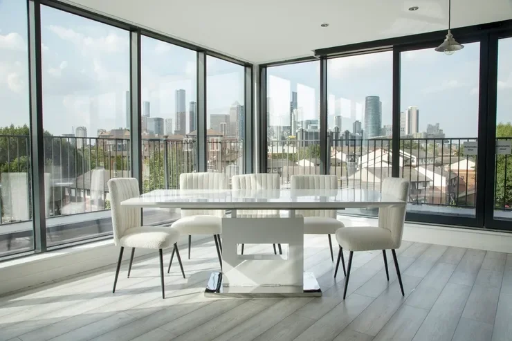 Empty dining table with eight white chairs in a modern apartment with large floor-to-ceiling windows and a city skyline view.