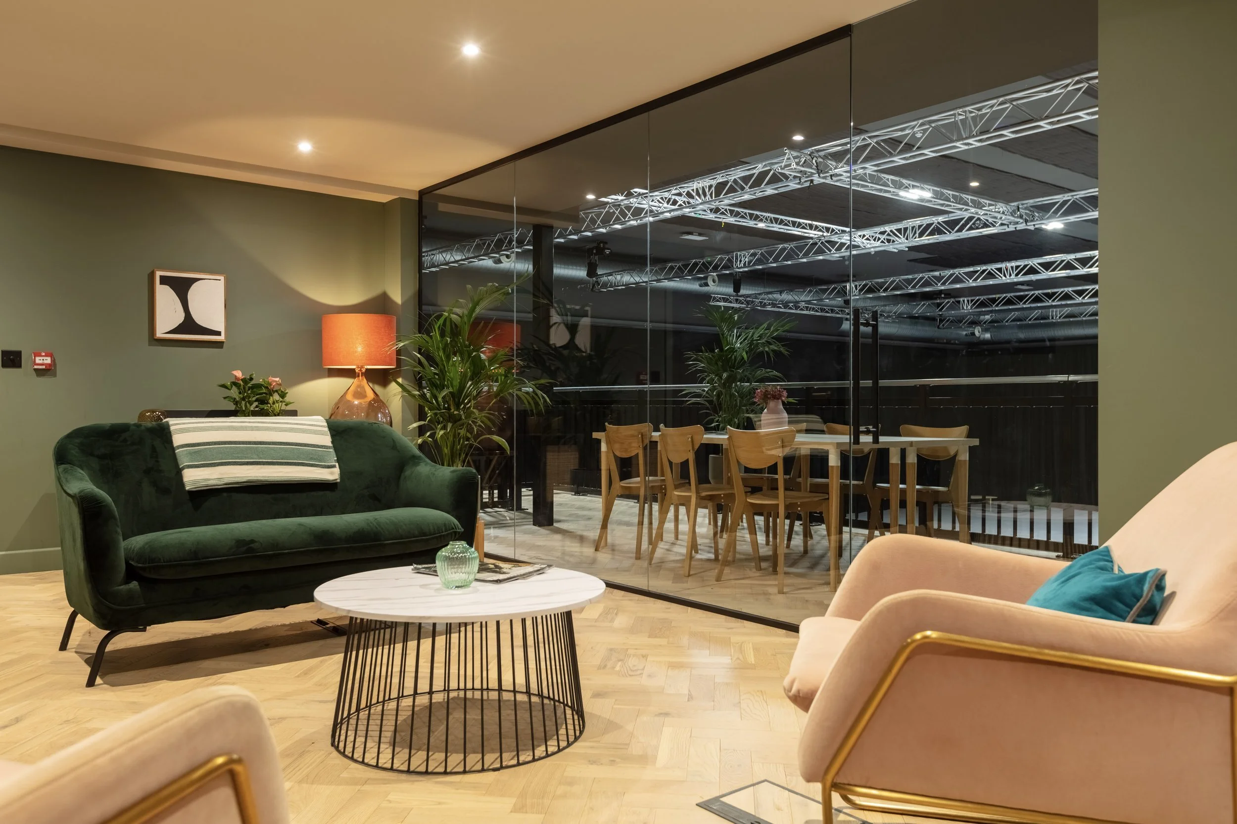 Interior of a modern living room with green and pink sofas, a round coffee table, indoor plants, a lamp, and a glass wall leading to a dining area with a wooden table and chairs.