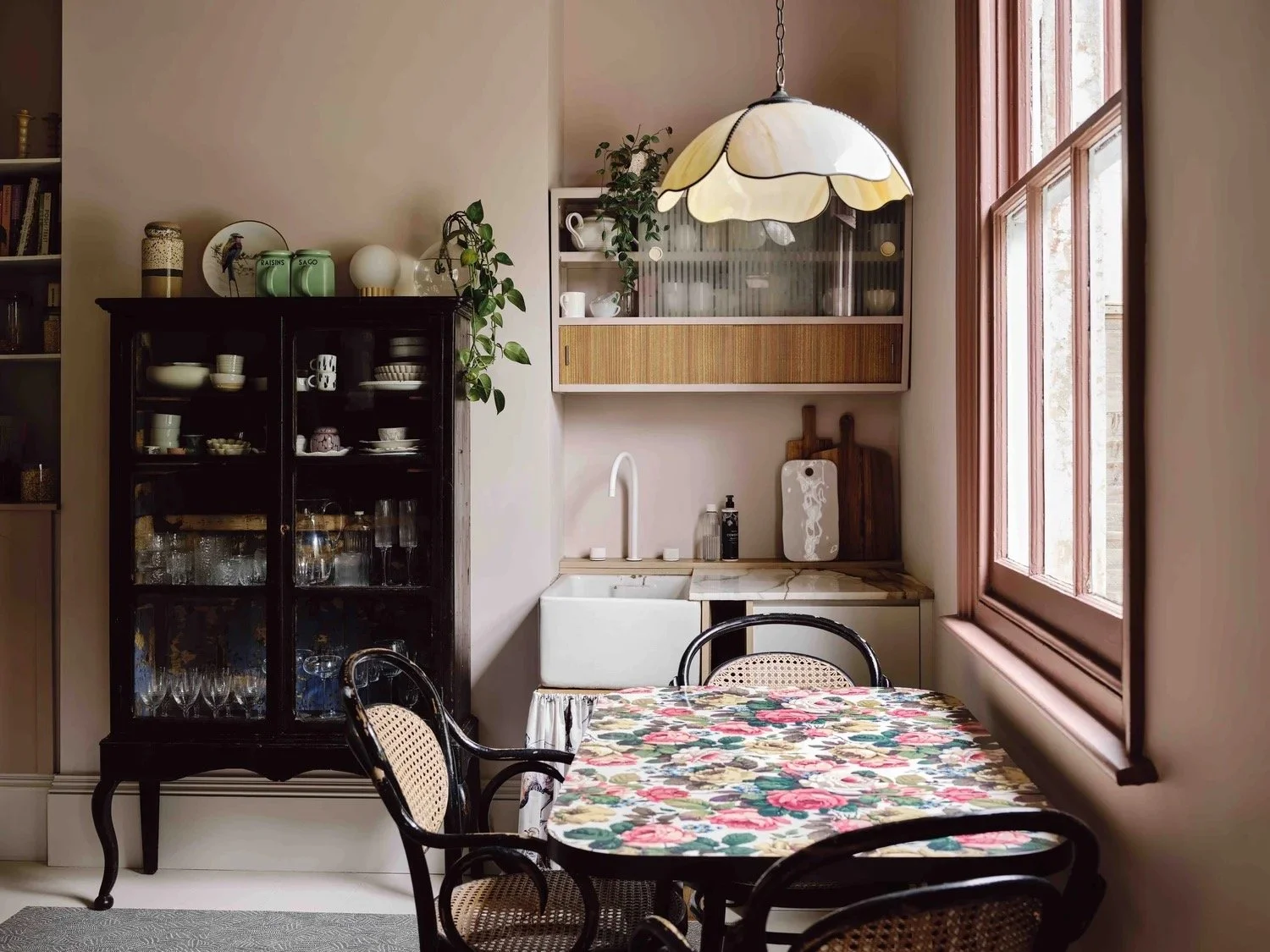 Cozy kitchen corner with a floral tablecloth-covered table, black chairs, a black glass-front cabinet displaying glassware and dishware, a wooden cabinet with a glass door, a marble countertop with a sink, a white wall-mounted cabinet, and a large wi