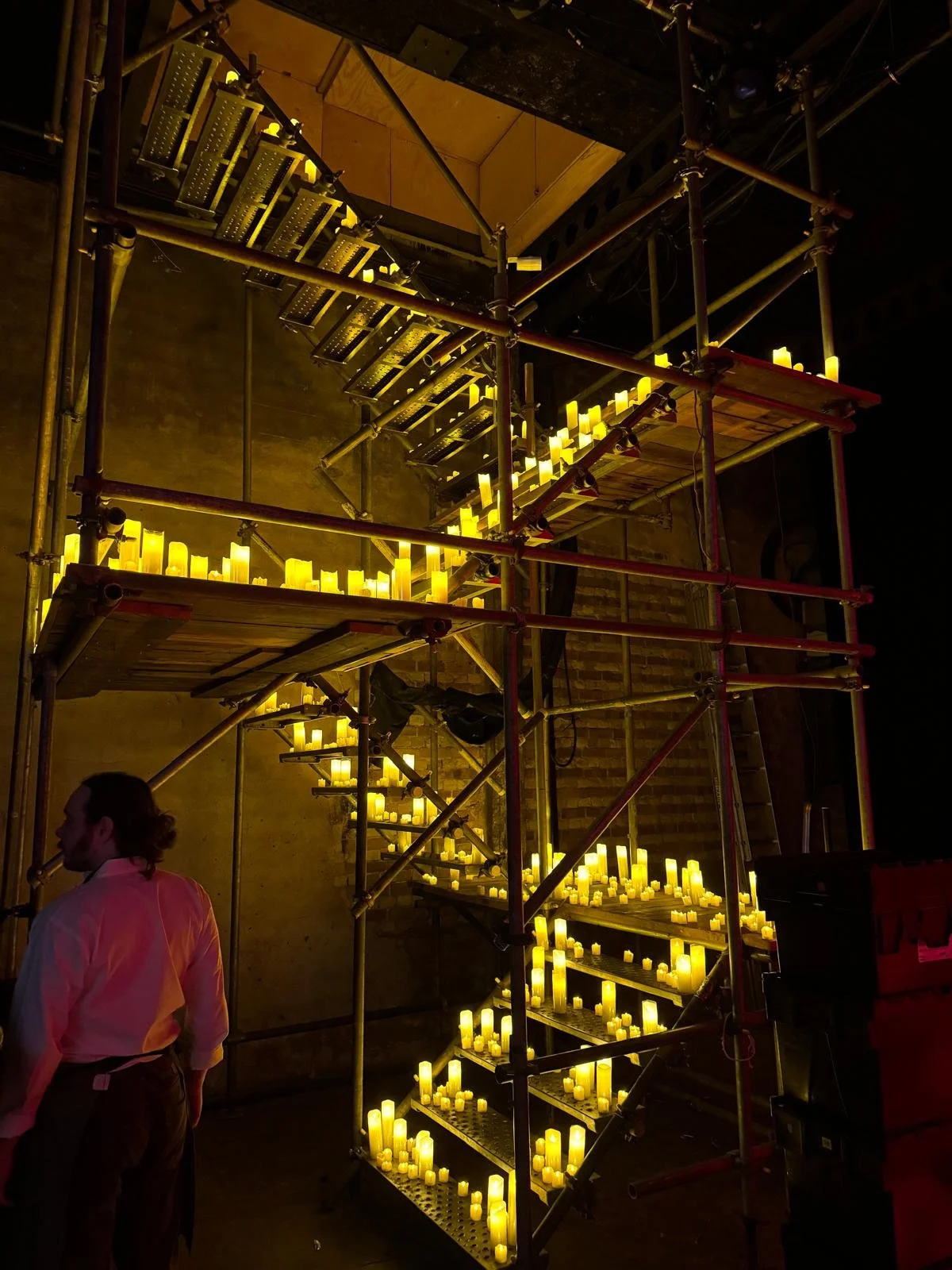 A multi-level scaffolding structure with numerous yellow candles on each platform, set against a dark background, with a person standing nearby.