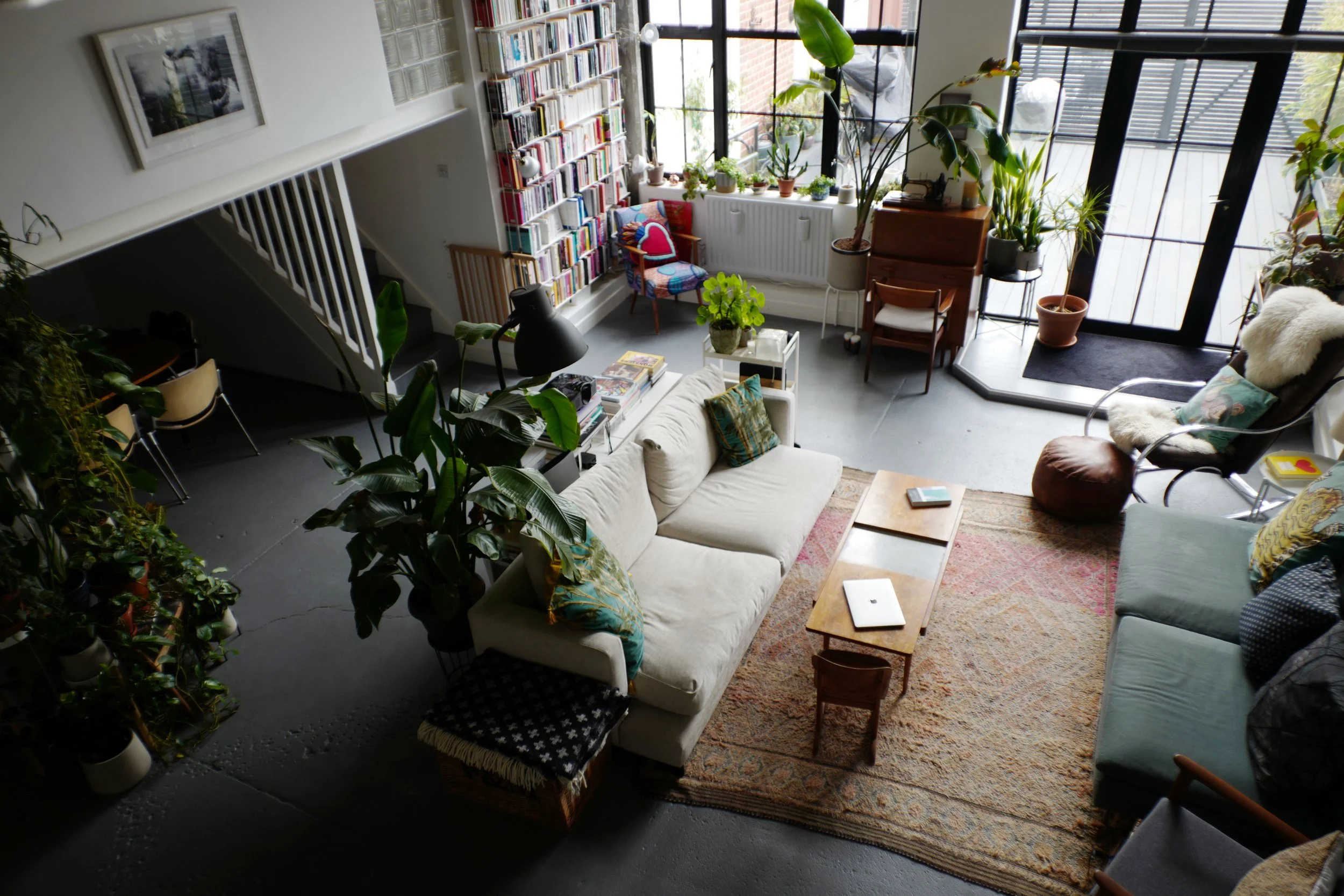 A cozy living room with large windows, a white sofa with colorful pillows, a wooden coffee table with a laptop and a book, surrounded by various potted plants, a green armchair with a fur throw, and a tall bookshelf filled with books.