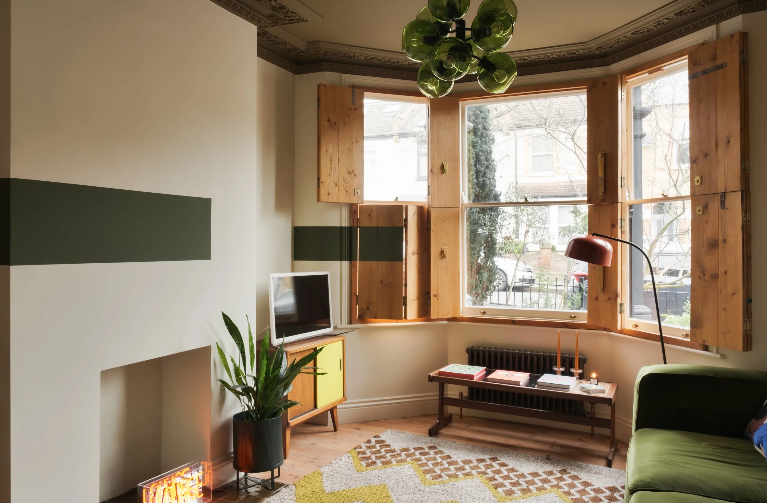 Living room with large bay window with open wooden shutters, green sofa, potted plant, wall-mounted TV, vintage coffee table with books, patterned rug, and modern chandelier with green glass globes.