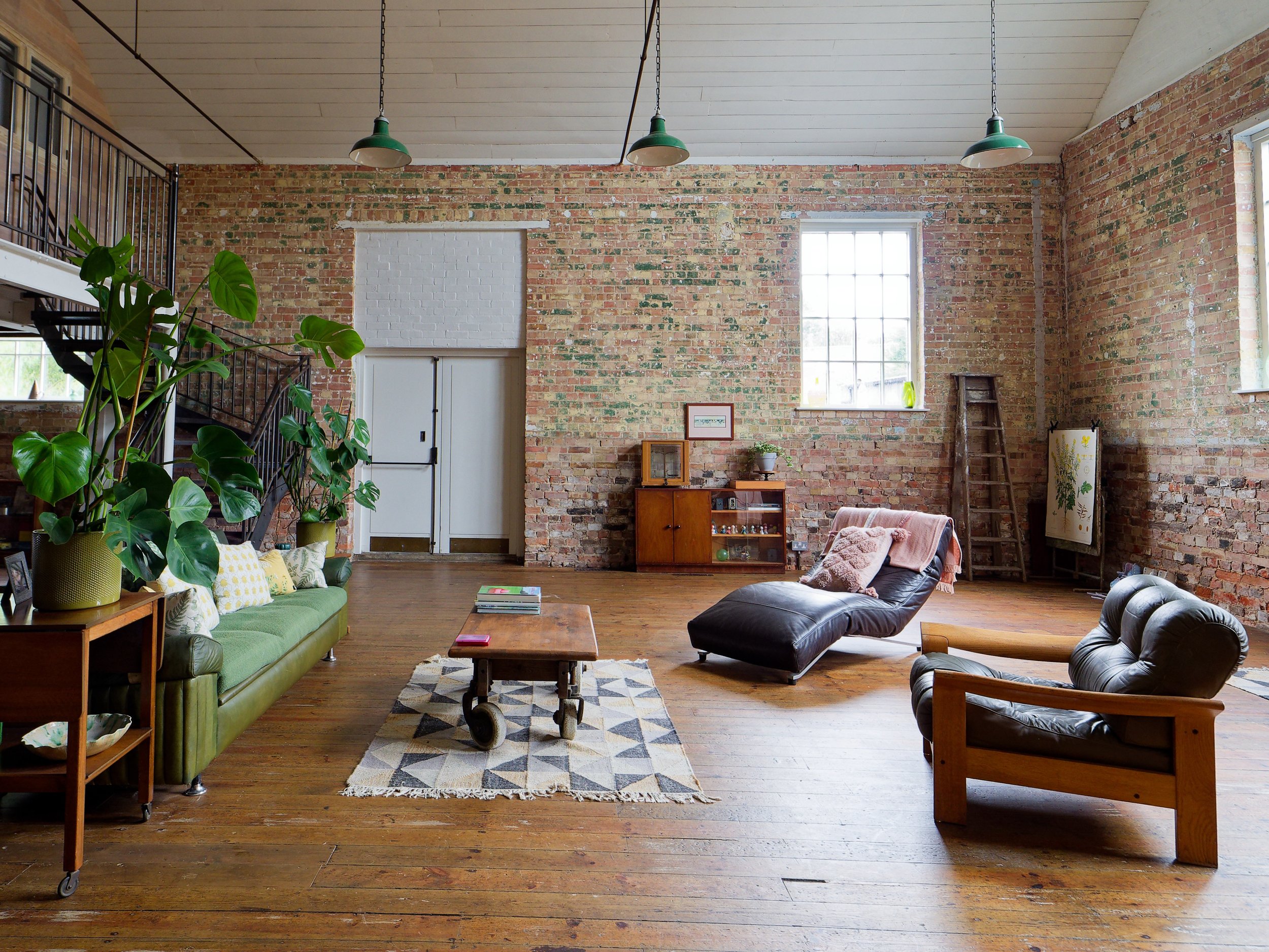 A spacious living room with brick walls, hardwood floors, and a high ceiling. There are large windows letting in natural light. The furniture includes a green sofa with yellow-patterned cushions, a black leather chaise lounge with a pink blanket and 