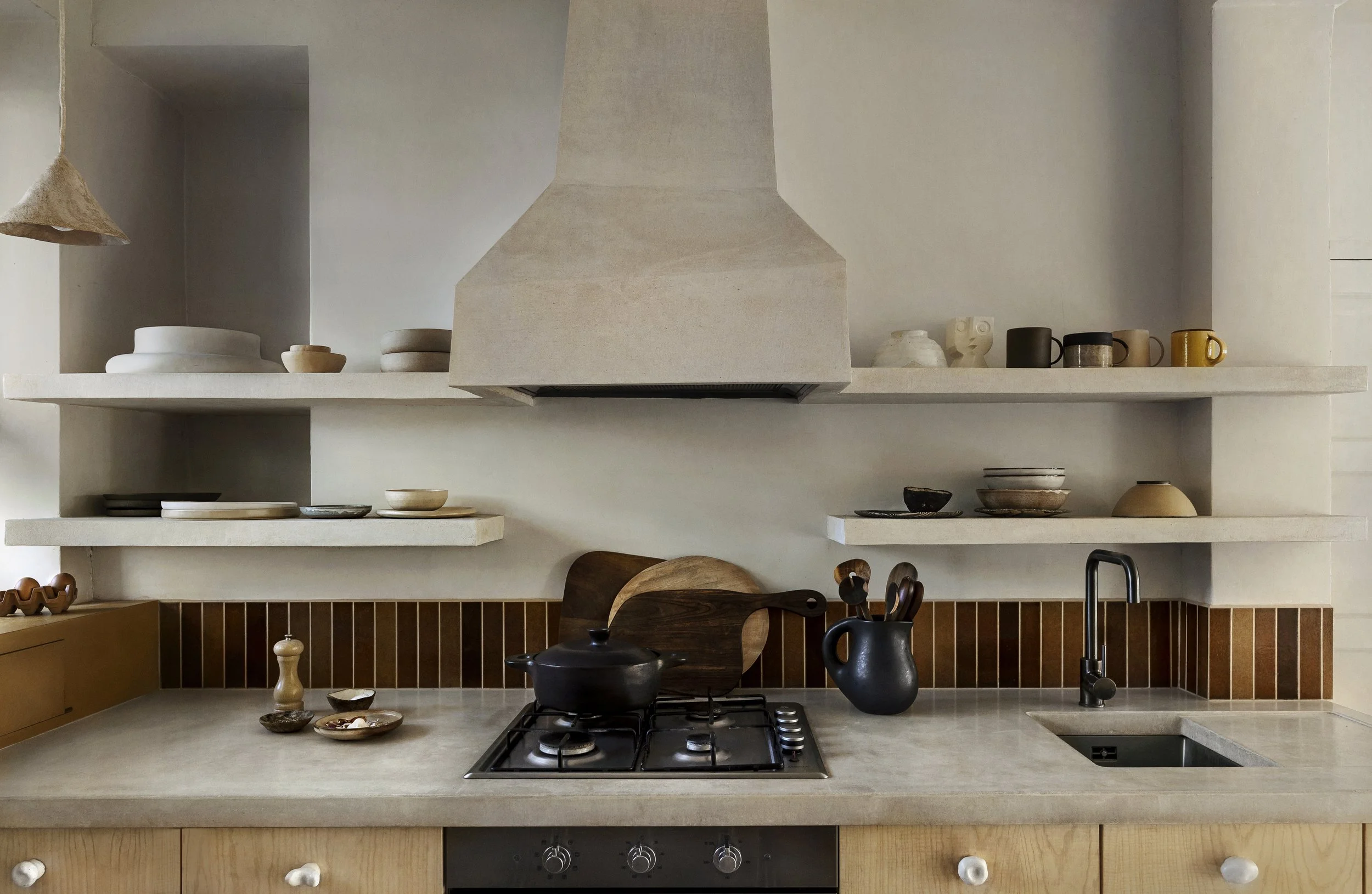 Minimalist kitchen with open shelves holding various dishes, a stove with a black pot, a black pitcher with utensils, and a small sink with a black faucet.
