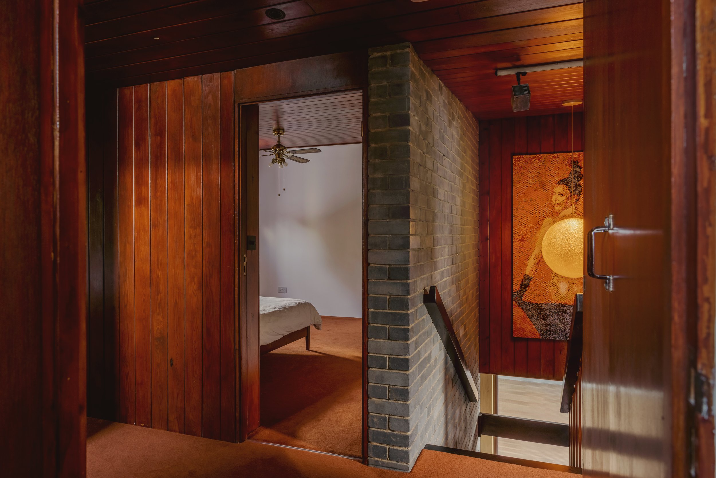 Interior view of a residential hallway with wood-paneled walls, a brick wall, a staircase, and part of a bedroom with a fan on the ceiling.