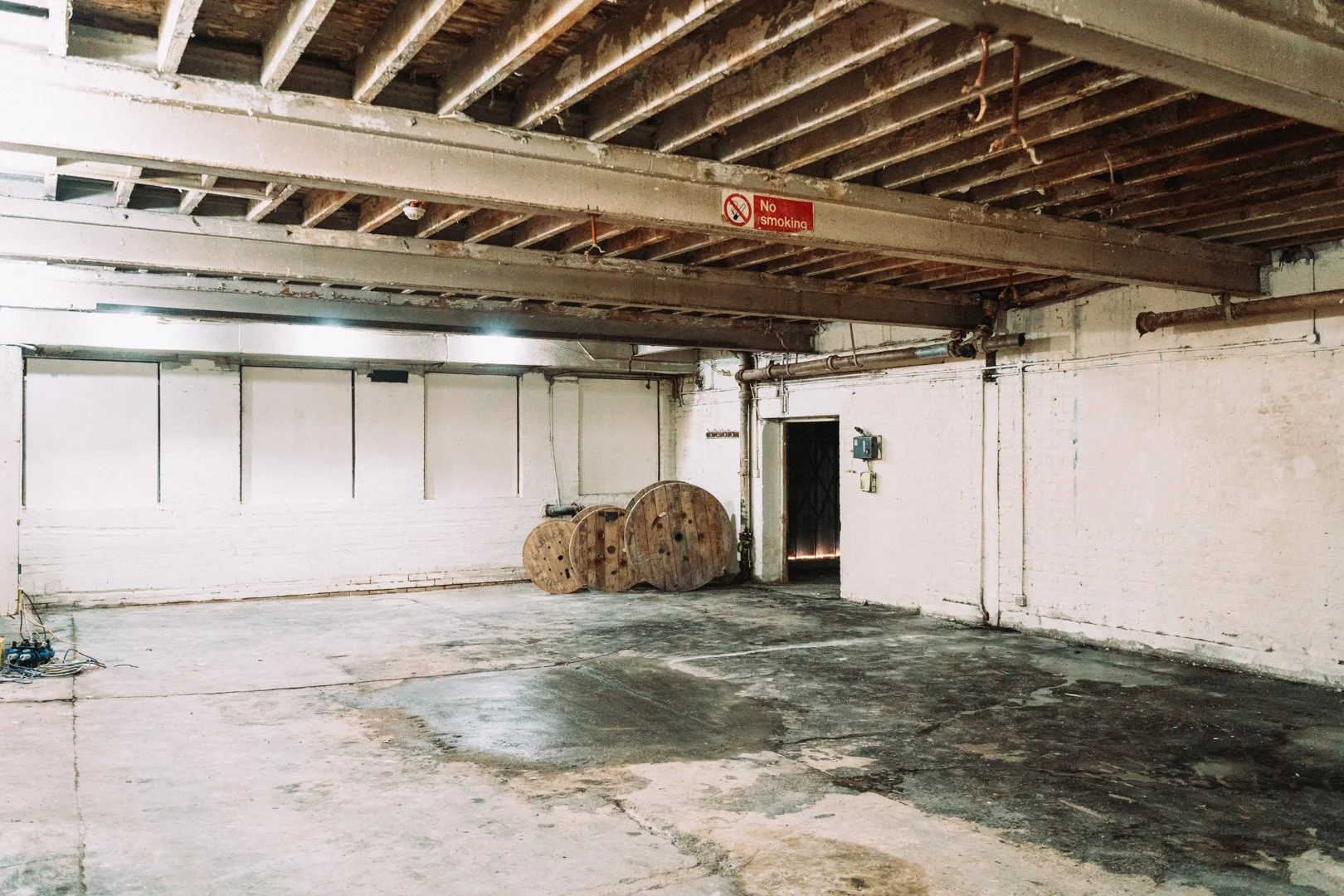 An empty industrial parking garage with unfinished ceiling beams, a 'No Smoking' sign, and three large wooden spools stacked near the back wall.