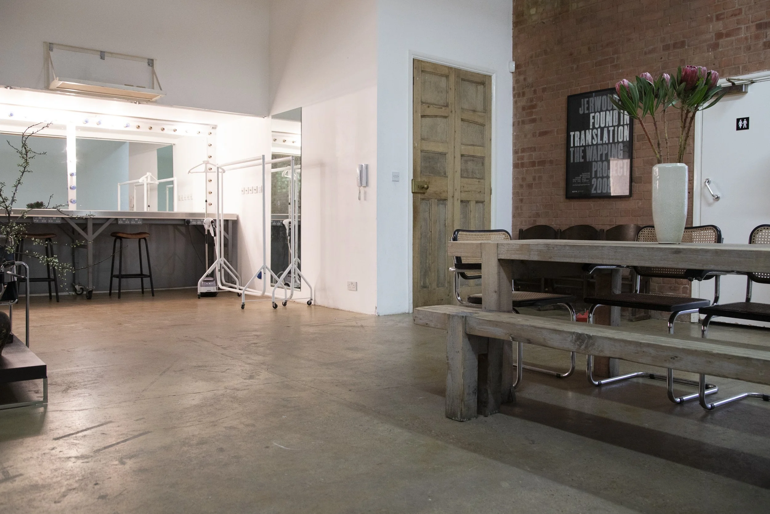 Interior of a modern cafe or workspace with a wooden bench, a table with a vase of pink flowers, chairs, a brick wall with a framed poster, and a white door, with a bar area visible in the background.