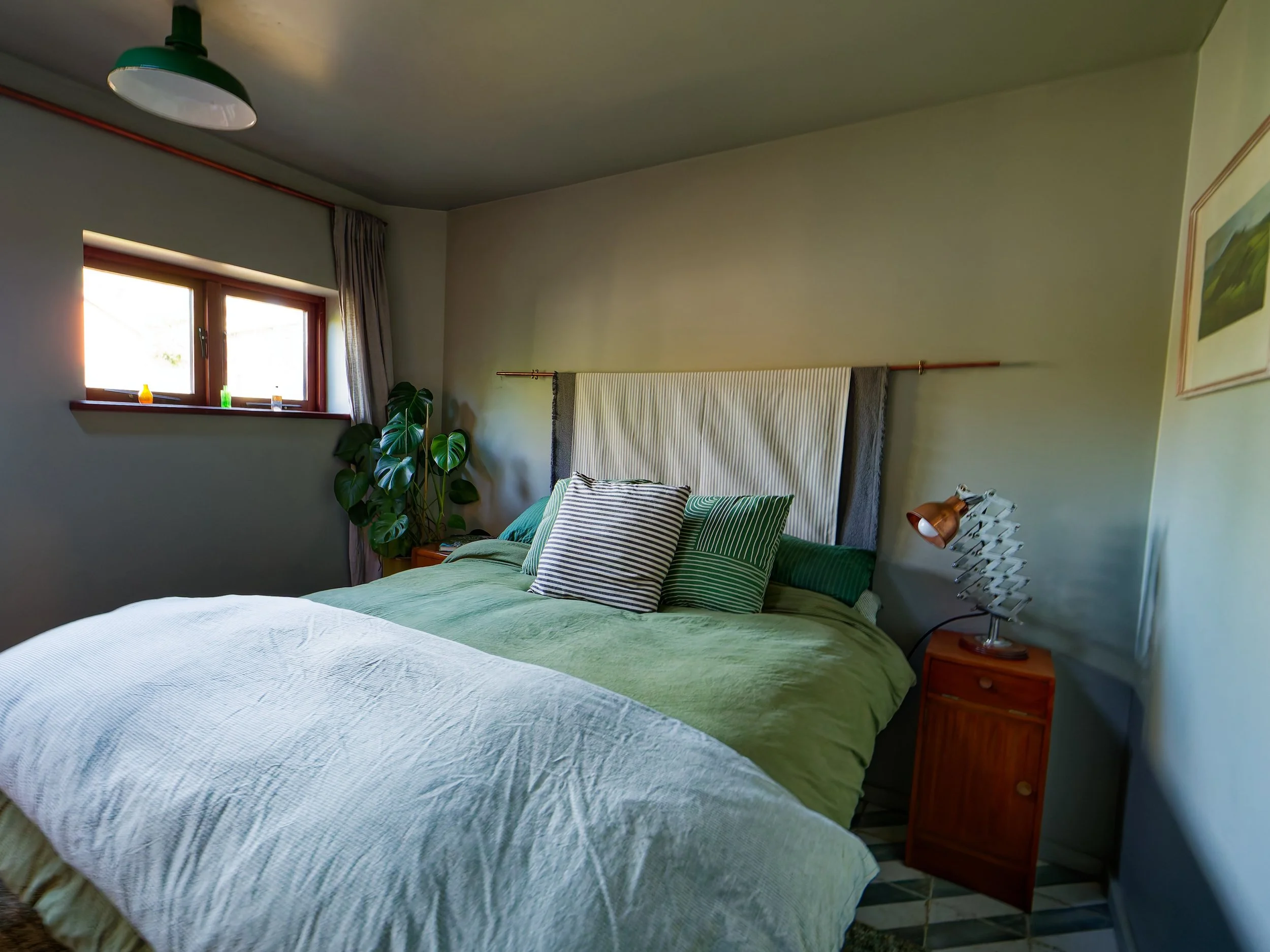 A cozy bedroom with a large bed dressed in green and white linens, decorative pillows, a wooden nightstand, a vintage-style desk lamp, a tall leafy plant near the window, and a framed landscape painting on the wall.