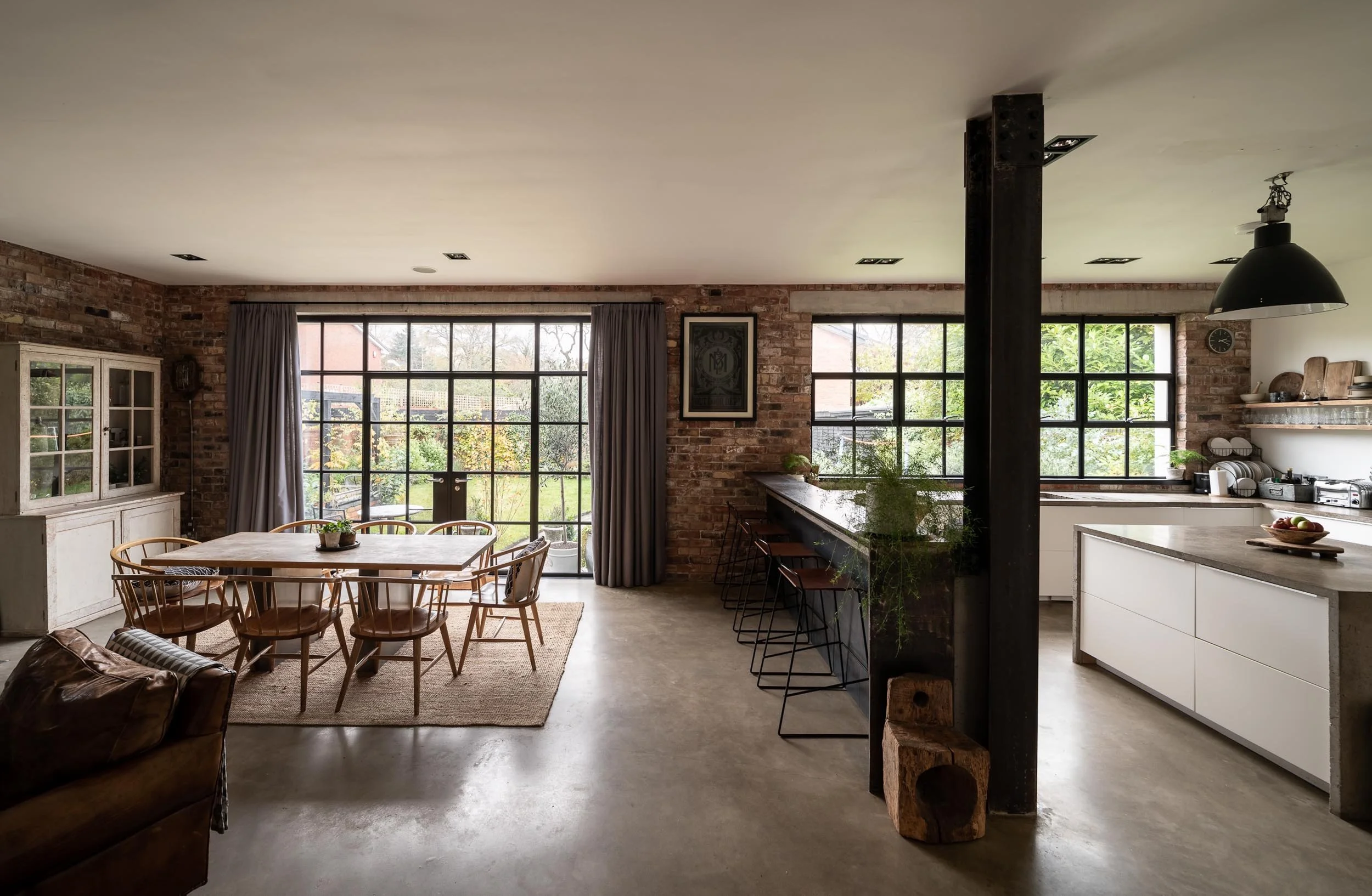 Open-concept living and dining area with brick walls, large sliding glass doors, wooden dining table with chairs, kitchen with white cabinetry, black bar stools, and outdoor garden view.