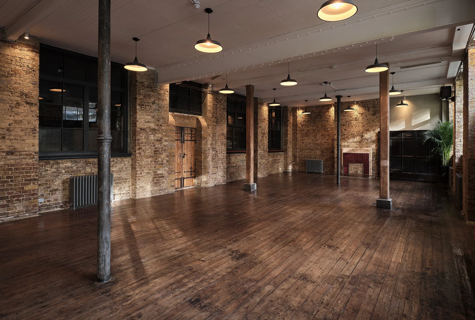 An empty industrial-style loft with exposed brick walls, wooden floors, black-framed windows, and ceiling lights.