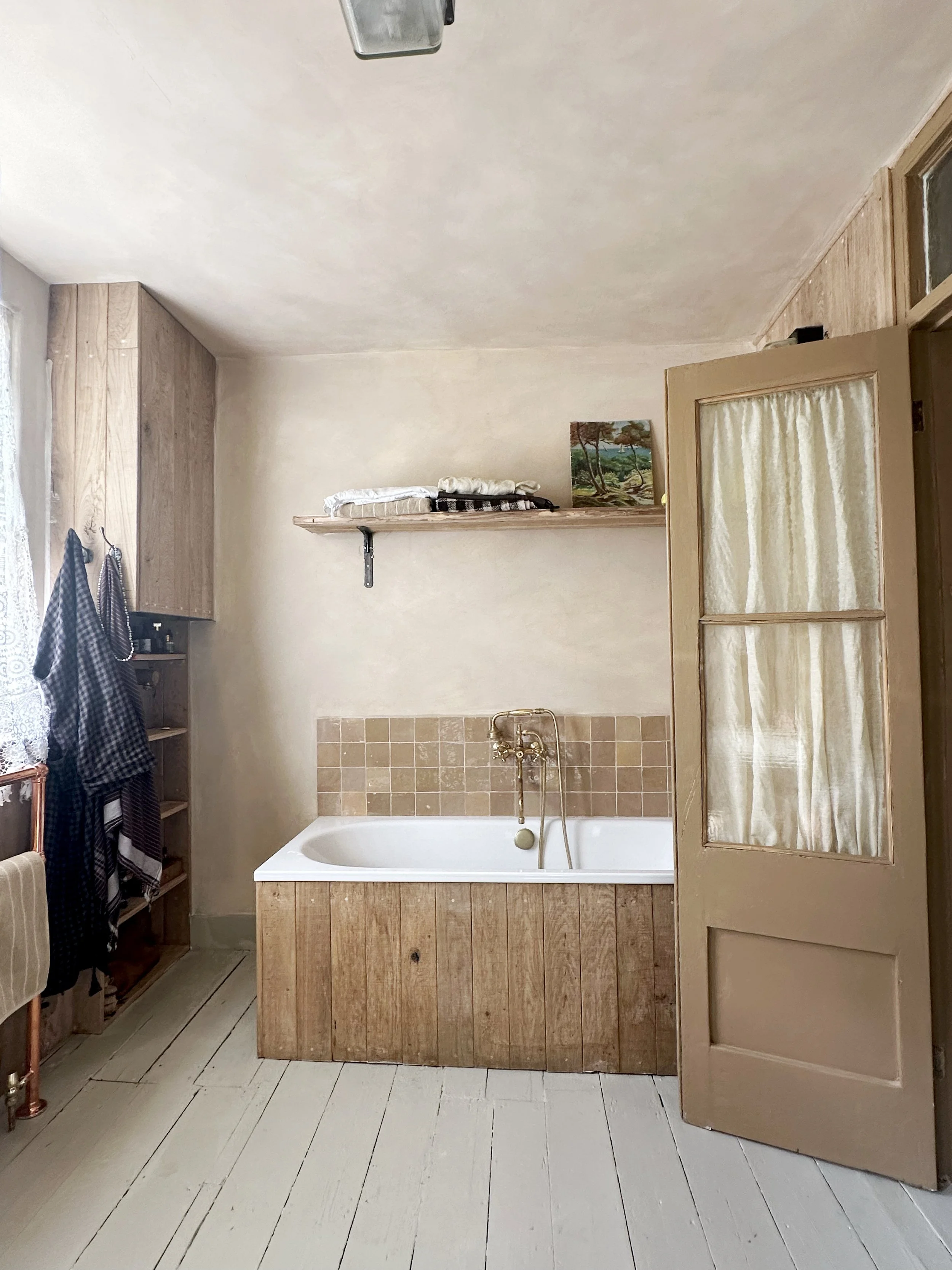 Bathroom with a bathtub, wooden paneling, a shelf with towels and a painting, a door with a curtain, and a window with lace curtains.