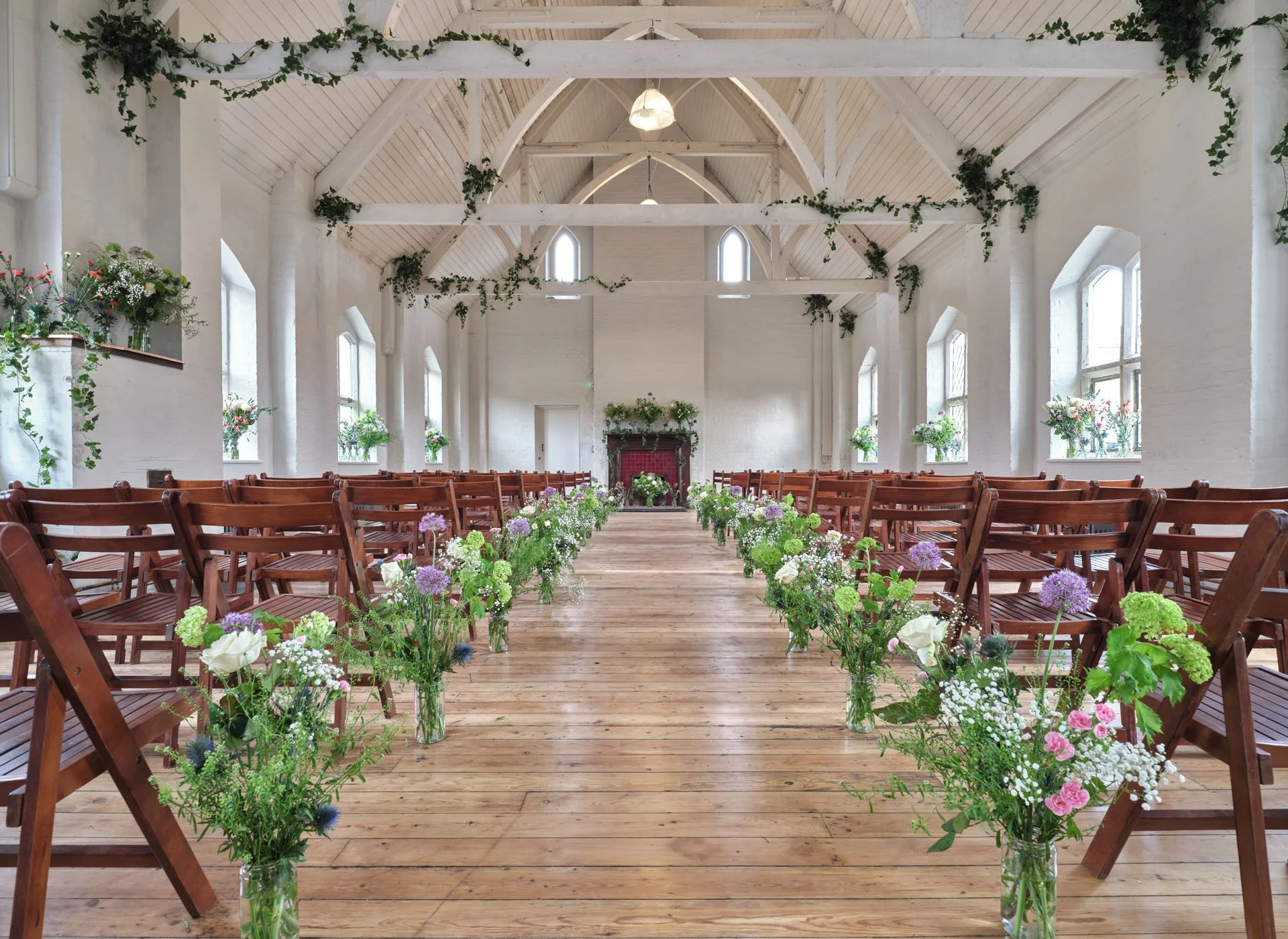 Wedding ceremony setup inside a bright, white chapel with wooden floors, wooden chairs arranged in rows, and glass vases filled with colorful flowers lining the aisle. The chapel has high, arched ceilings with exposed beams and multiple windows letti
