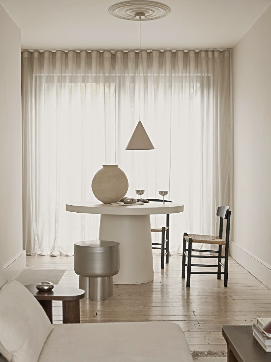 Minimalist dining and living room with white curtains, a round white table, black chairs, a gray stool, and a beige sofa.
