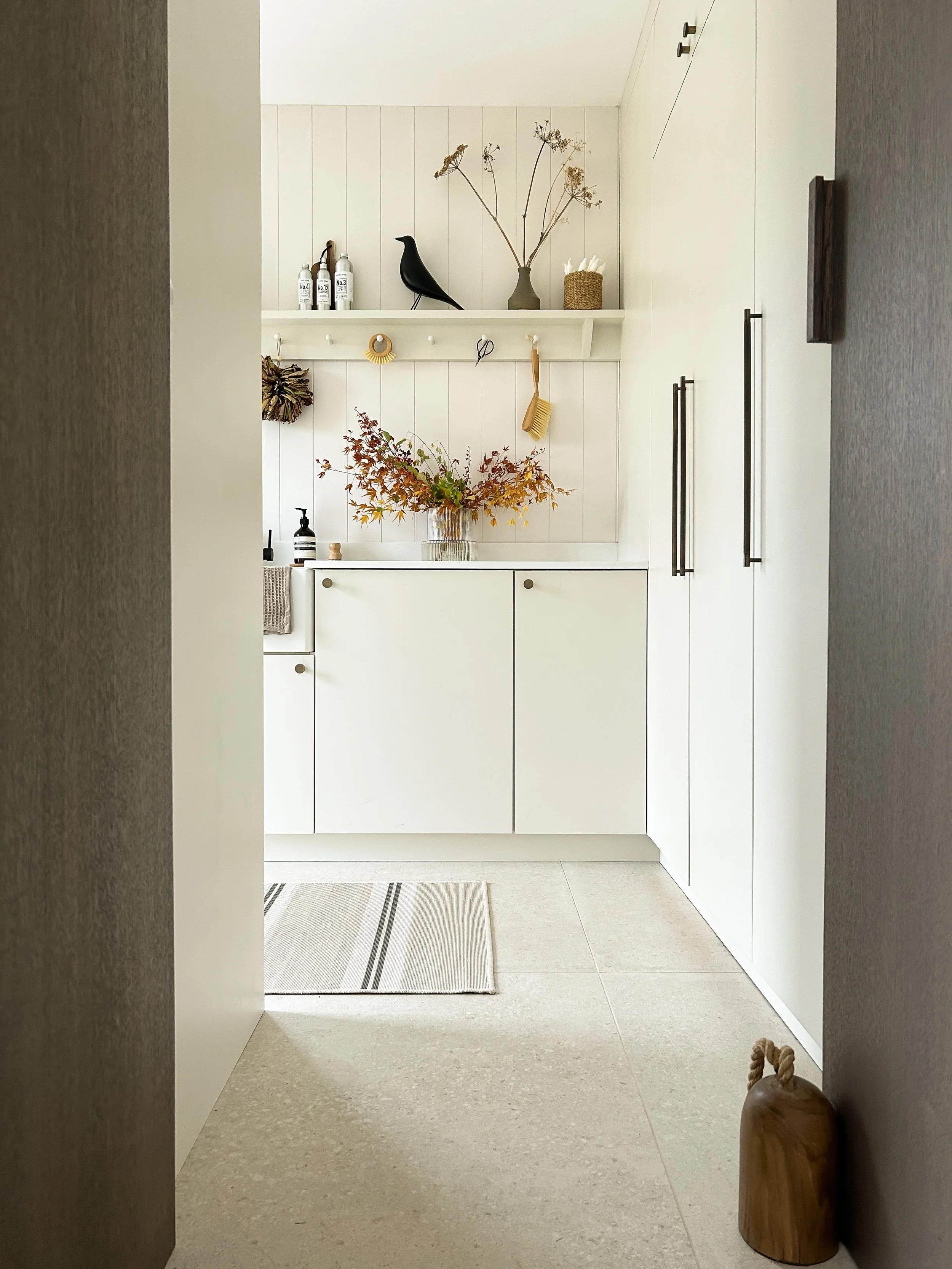 A minimalist bathroom with white cabinetry, a vase of autumn leaves on the counter, decorative items on the shelves, and a small mat on the floor.