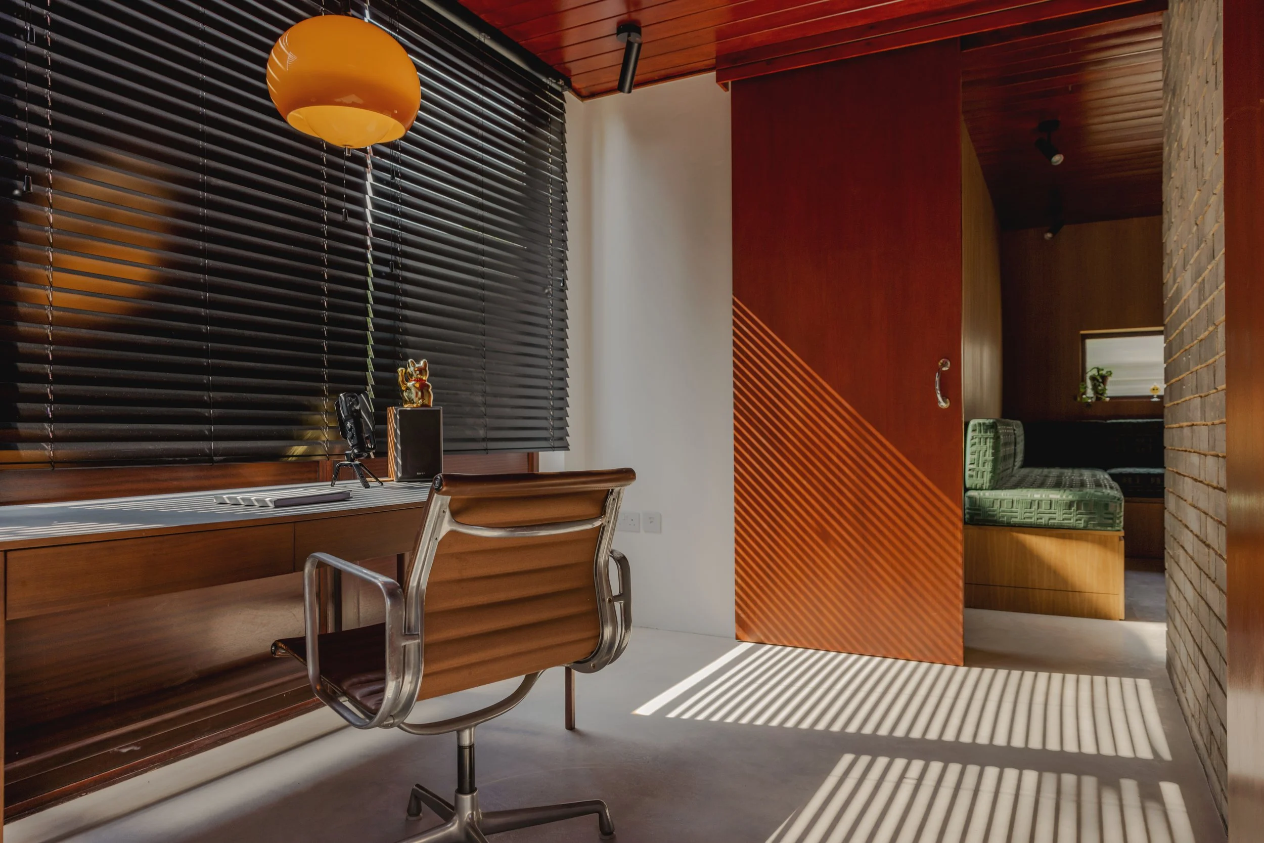 Modern office with wooden desk, beige swivel chair, dark blinds on window, orange hanging lamp, and sunlight creating striped shadows on the floor; partially open sliding door revealing a cozy green upholstered seating area inside a room.