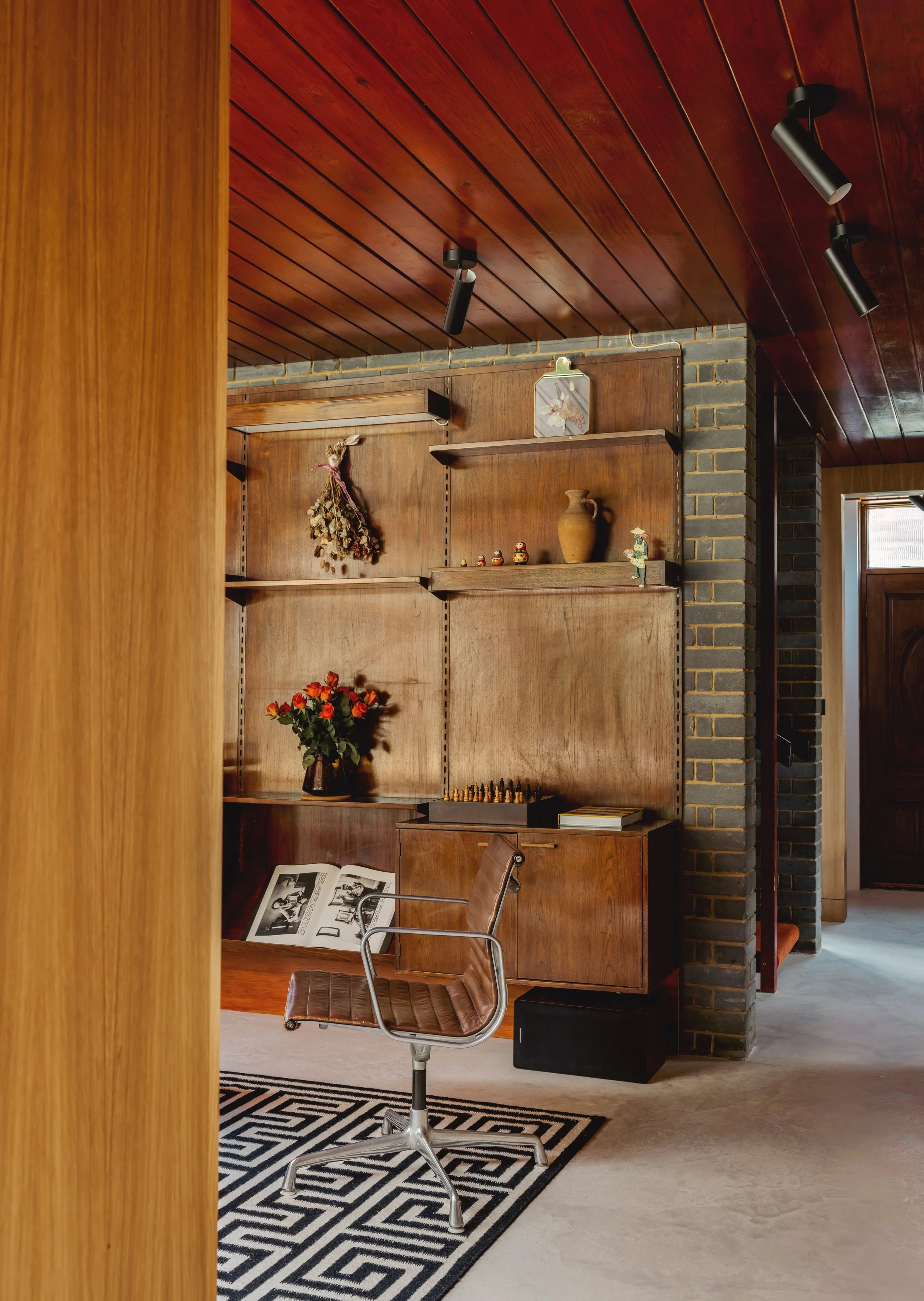 A mid-century modern home office with wood-paneled walls and ceiling, a vintage brown leather office chair, a wooden desk with an open magazine, a black and white geometric rug, and decorative items including a vase of orange flowers, shelves with dr