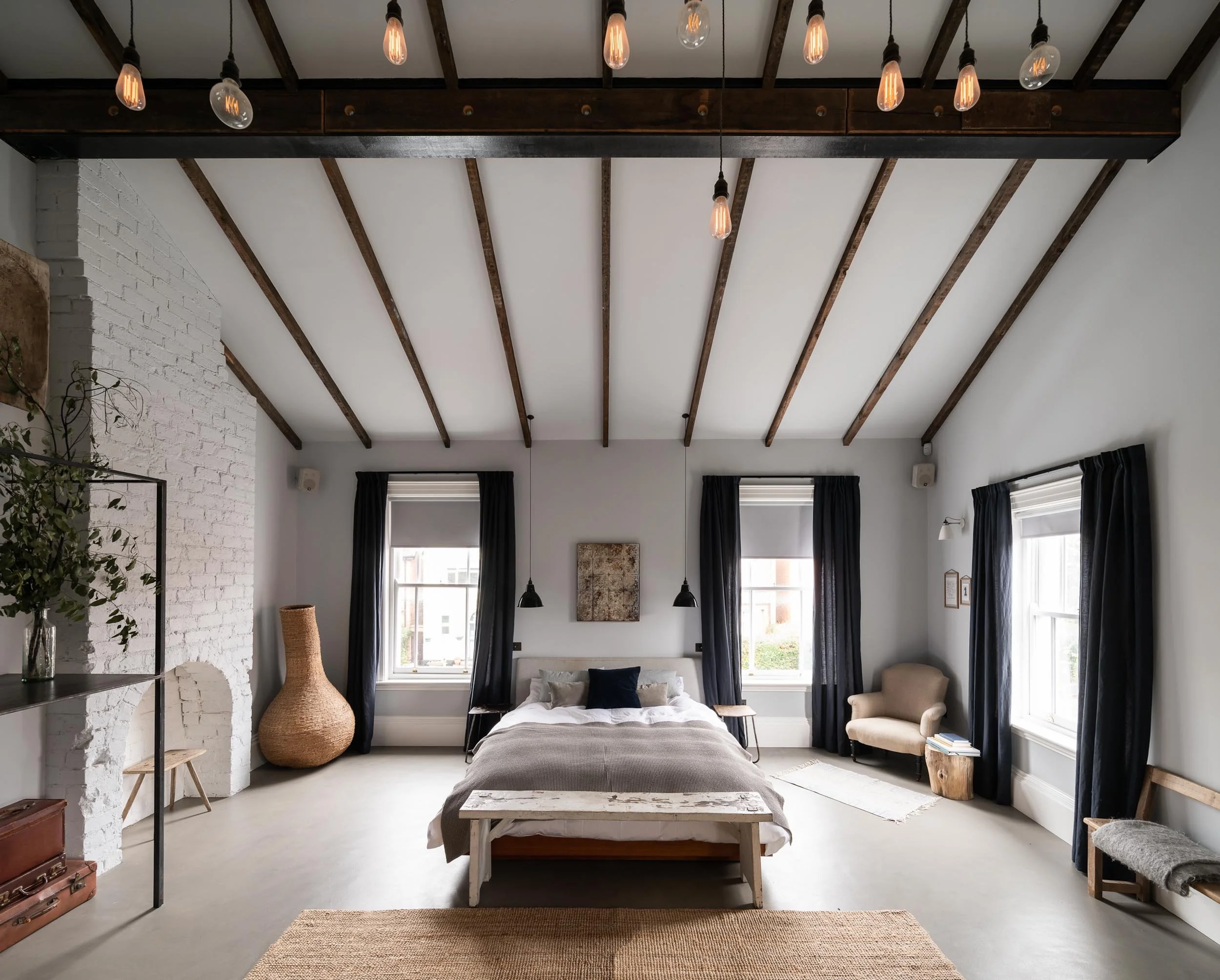 A spacious bedroom with white walls and ceiling, wooden beams, and three large windows with black curtains. There is a bed with neutral bedding, a small side table, and various decorative items including a wicker vase, a framed artwork, and a beige a