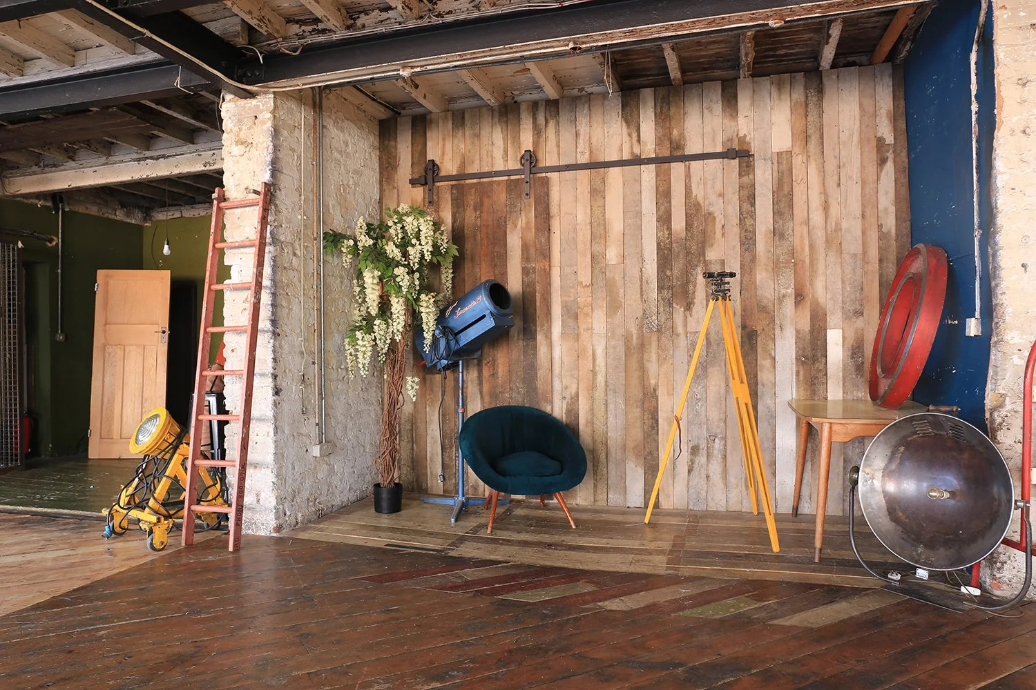 Interior space with wooden floor and wall, a red ladder, a yellow work light, a potted plant with white flowers, a blue vacuum, a dark green upholstered chair, a yellow tripod, a small wooden table, a large red mirror, and a large metallic reflector.