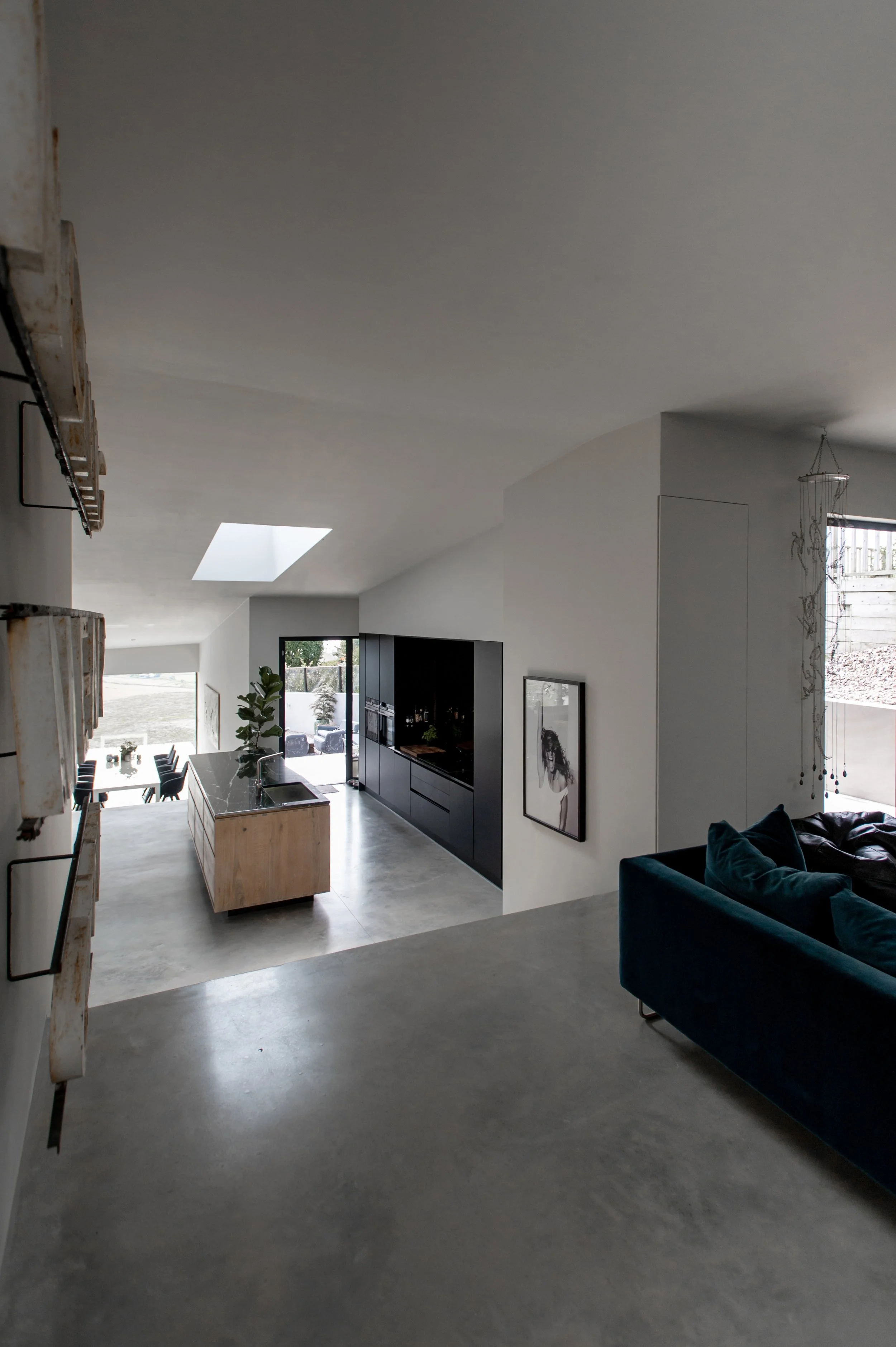 Interior view of a modern, open-concept living space featuring a kitchen with a wooden island, black cabinetry, and a gray concrete floor. There's a dark blue sofa on the right and artwork on the wall. A sliding glass door leads to an outdoor area wi