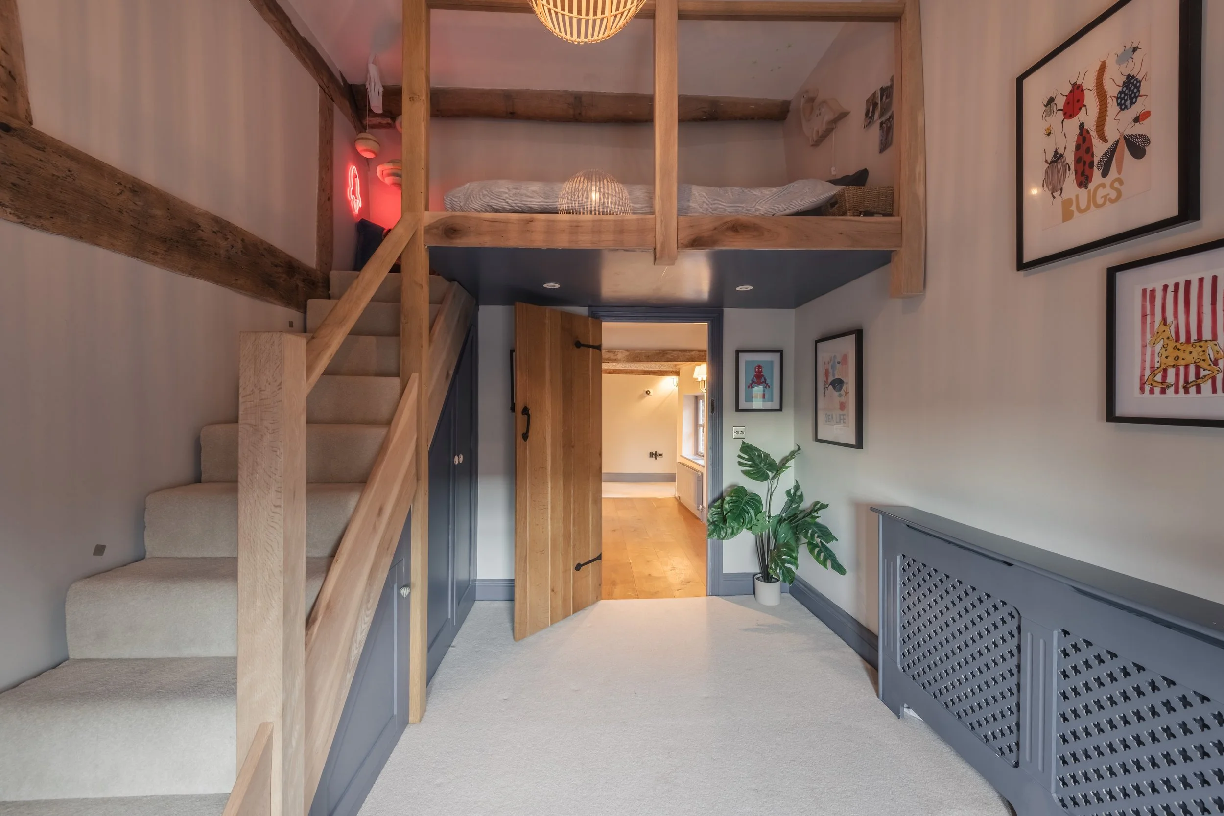 Interior of a cozy home with a staircase leading to a loft bedroom. The room features wood accents, framed colorful illustrations on the wall, a potted plant, and a door opening to another room with hardwood floors.
