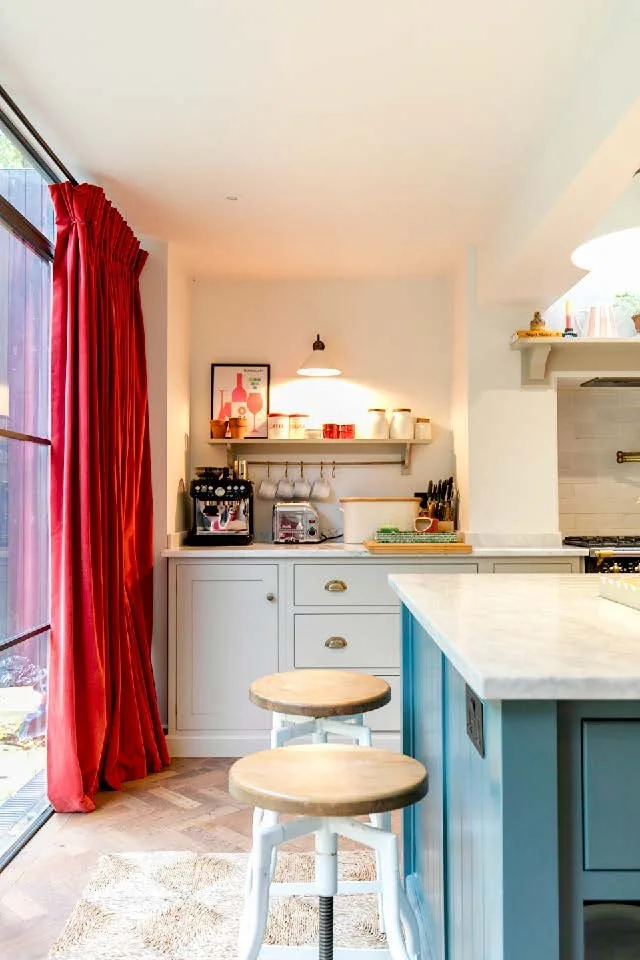 A cozy kitchen corner with a blue island, two wooden bar stools, a white cabinet, shelves with jars and artwork, red curtains, and a coffee maker near a glass sliding door.