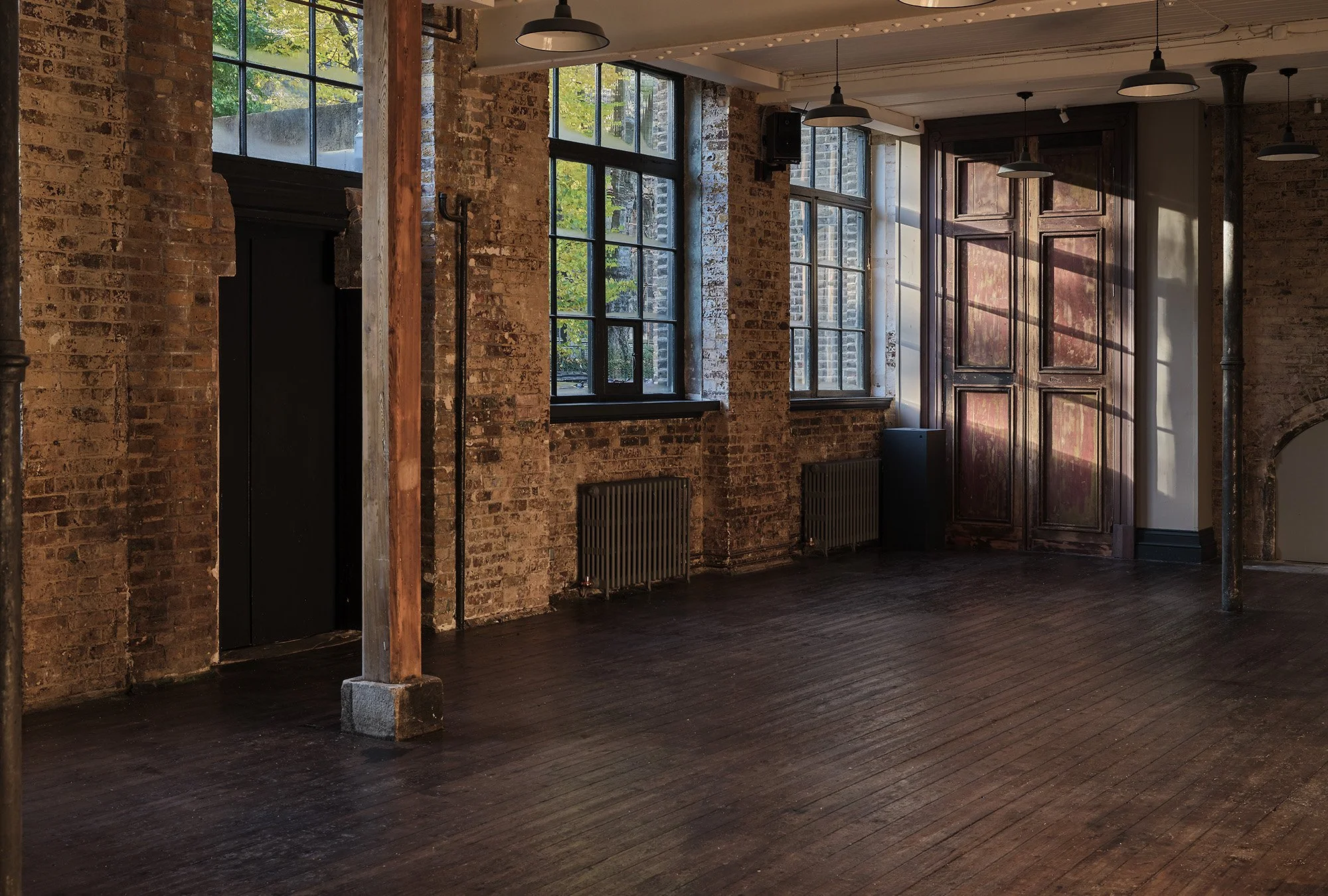 An empty industrial-style interior space with exposed brick walls, large windows, dark wooden flooring, black radiators, and ceiling-mounted light fixtures. There are supports and a wooden door with sunlight casting shadows inside.