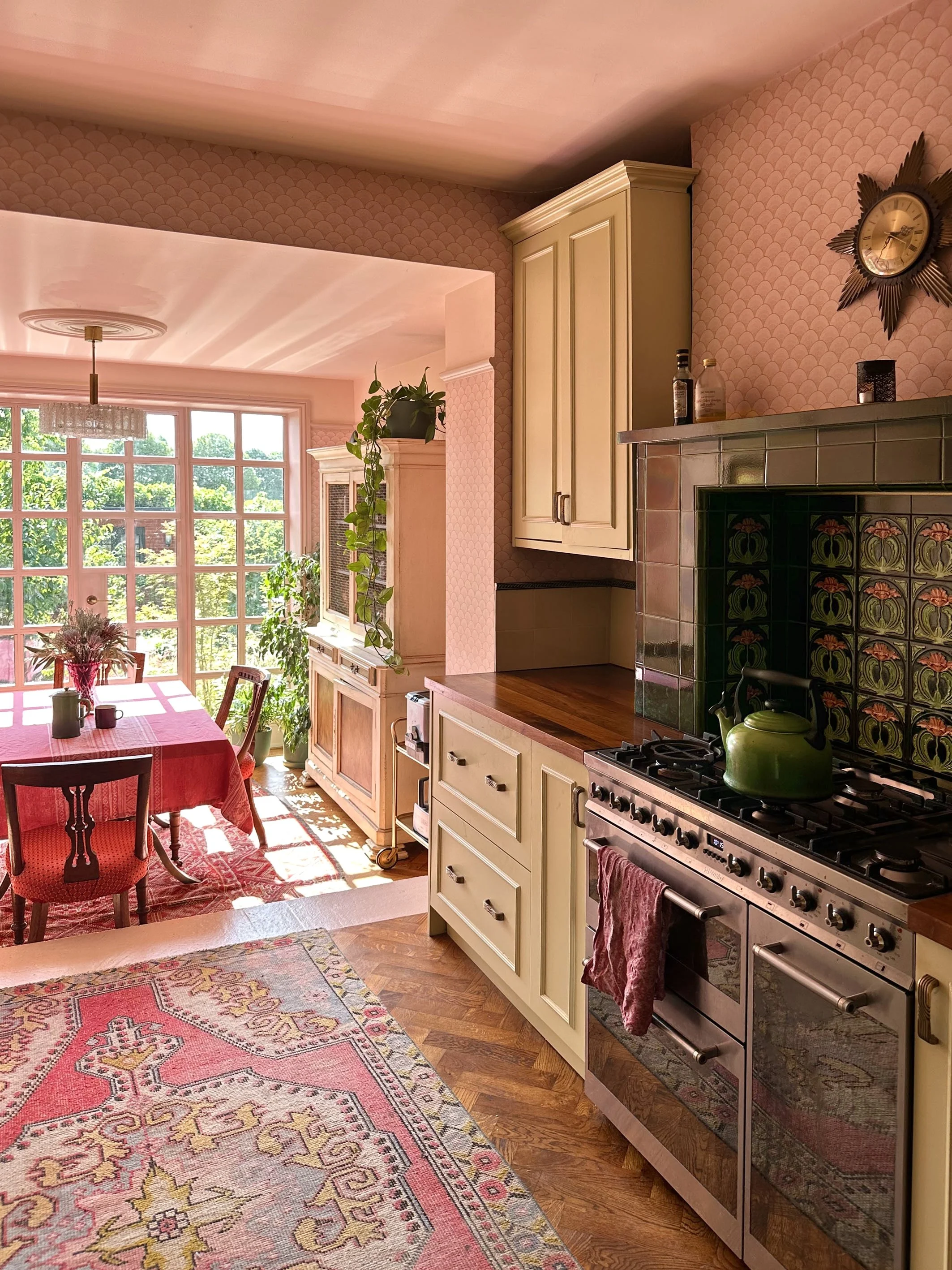 Kitchen with vintage green stove, cream-colored cabinets, pink patterned wallpaper, pink rug, dining table with red tablecloth, large windows with sunlight, and indoor plants.