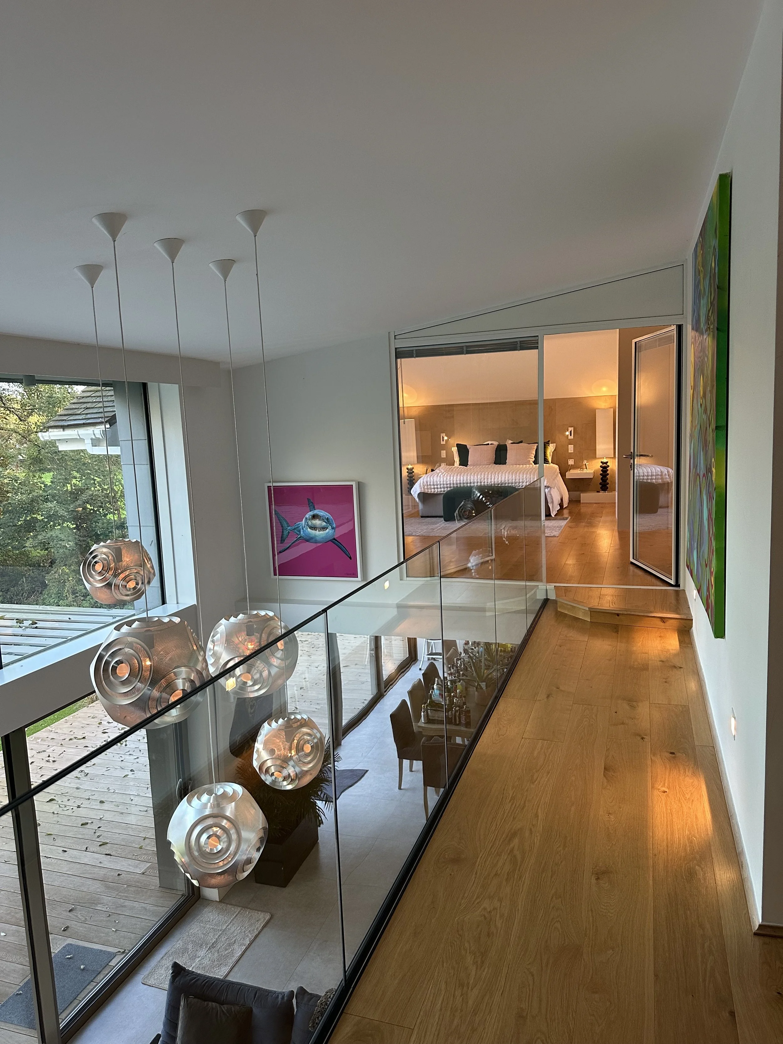 Interior view of a modern house with a staircase leading to a bedroom, featuring large windows, hanging pendant lights, colorful artwork, and a view of a patio outside.
