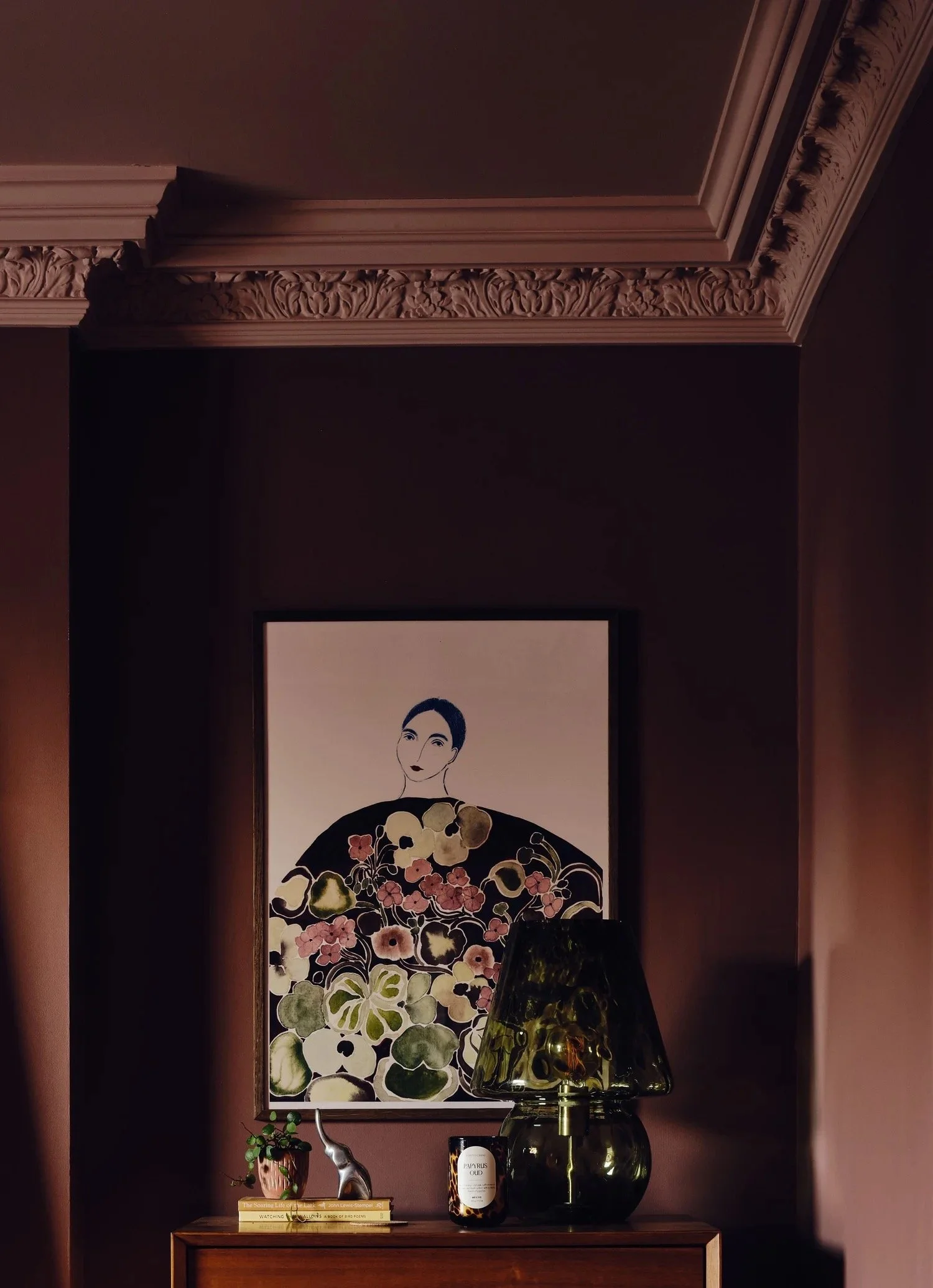 Decorative interior with a framed artwork of a woman with dark hair and floral dress, a small potted plant, a candle, and a glass lamp on a wooden console against a brown wall.