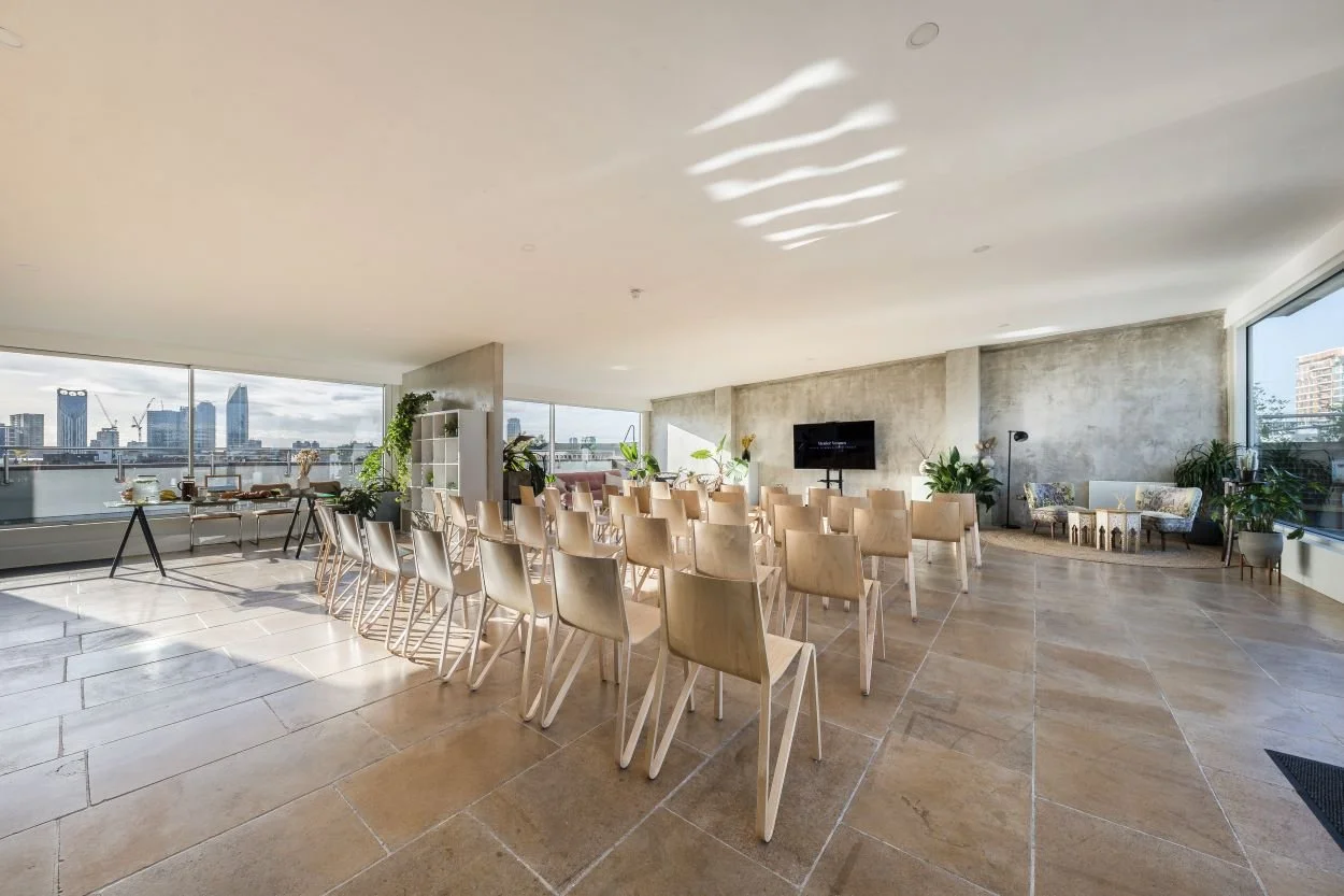A spacious, modern conference room with rows of wooden chairs facing a large flat-screen TV on a concrete wall, large windows with cityscape views, and abundant potted plants.