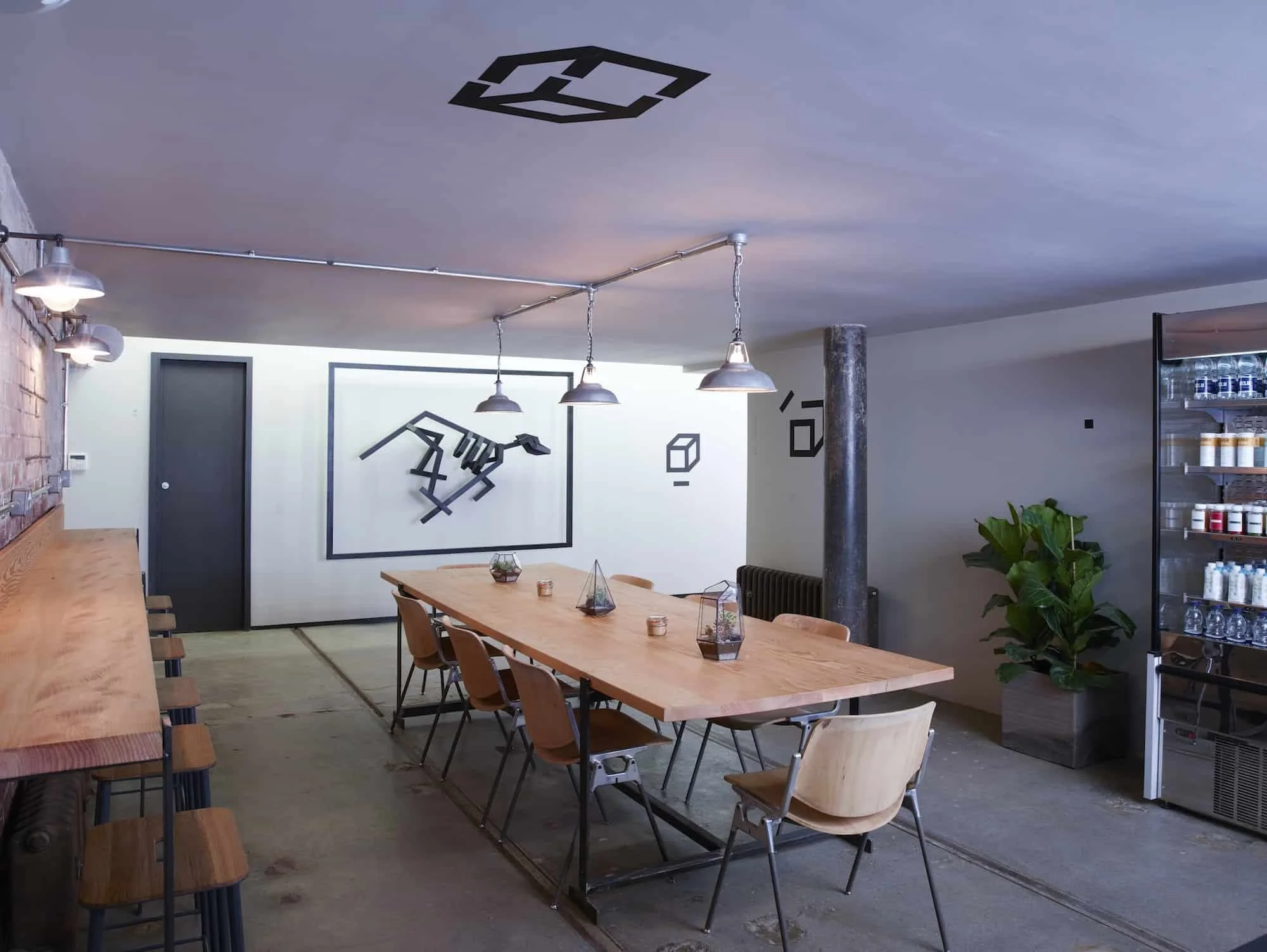 Modern dining room with a long wooden table, wooden chairs, and a wall-mounted artwork of a dog's outline made of black lines. There are pendant lights hanging above the table, a potted plant in the corner, and a refrigerated display case with bottle