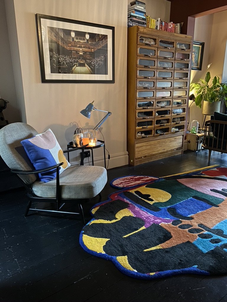 A cozy living room corner with a beige armchair, colorful pillow, small side table with a desk lamp and candle, a multicolored abstract rug, a wooden cabinet with multiple small drawers, and a large plant in a yellow pot.