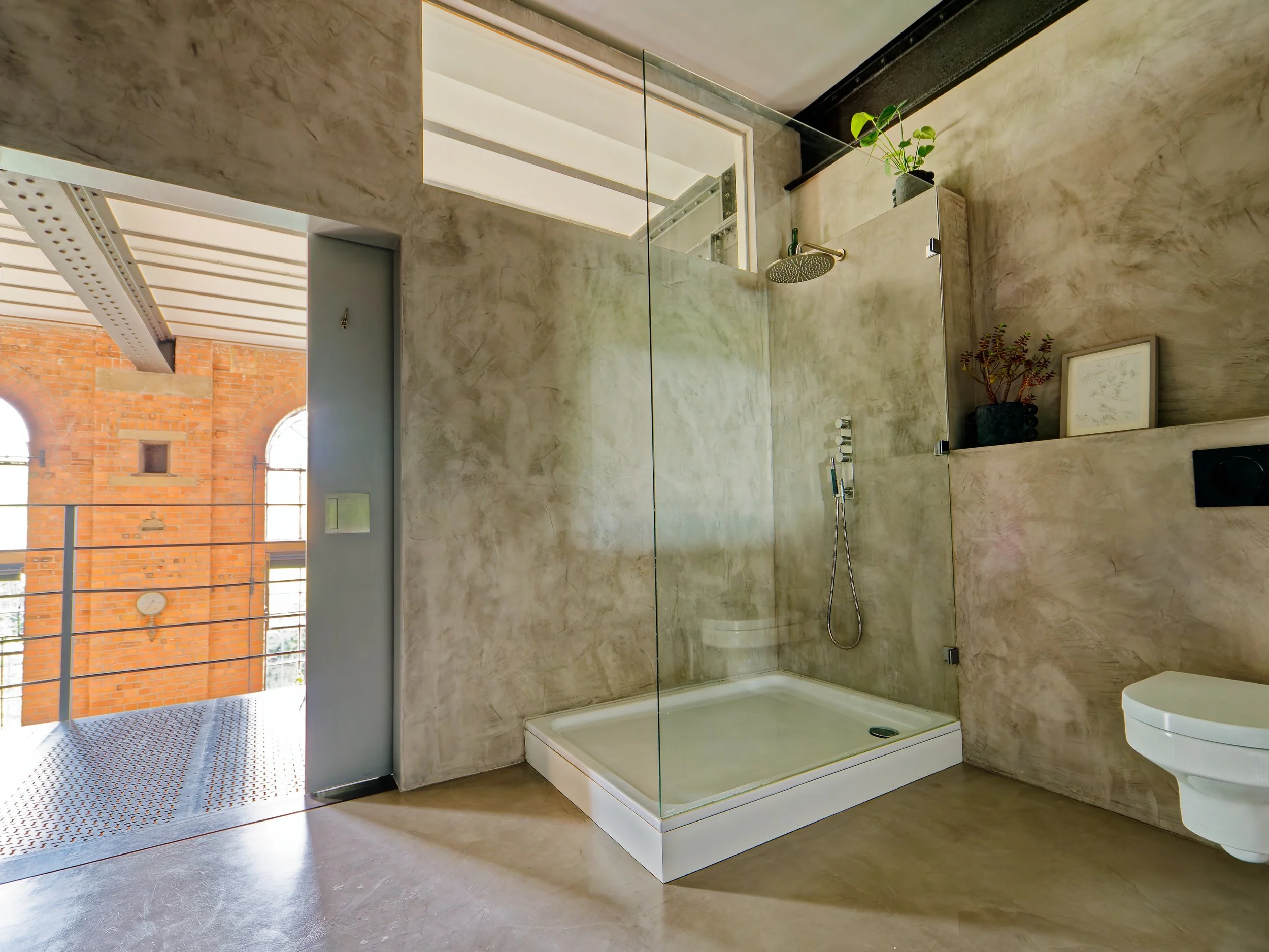Modern bathroom with concrete walls, a glass-enclosed shower area, a small balcony with a brick building outside, and decorative plants.