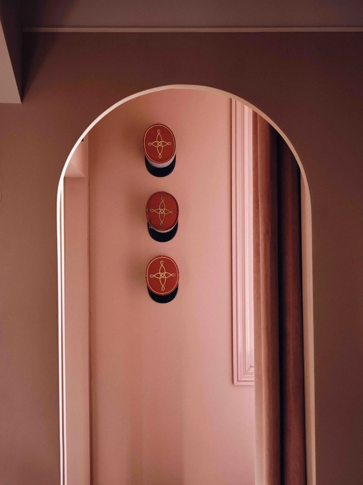 Three decorative wall plaques with a red background and gold design, mounted vertically on a pink wall next to a window with beige curtains.