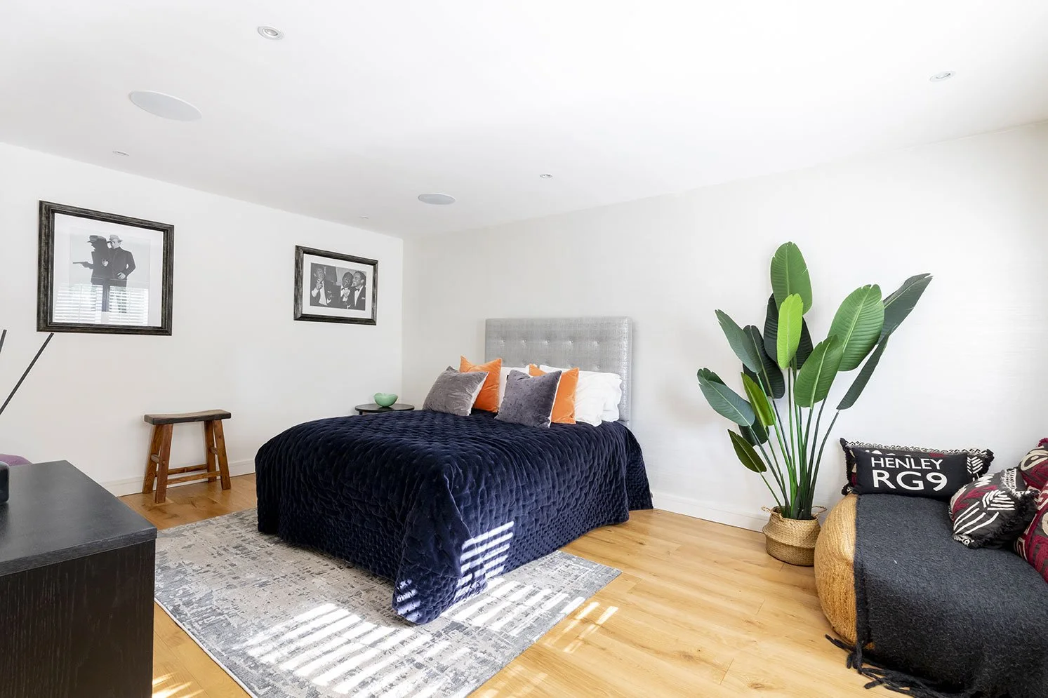 Bedroom with a bed covered in a dark blue quilt, multiple pillows, a large potted plant, and framed pictures on the white wall.