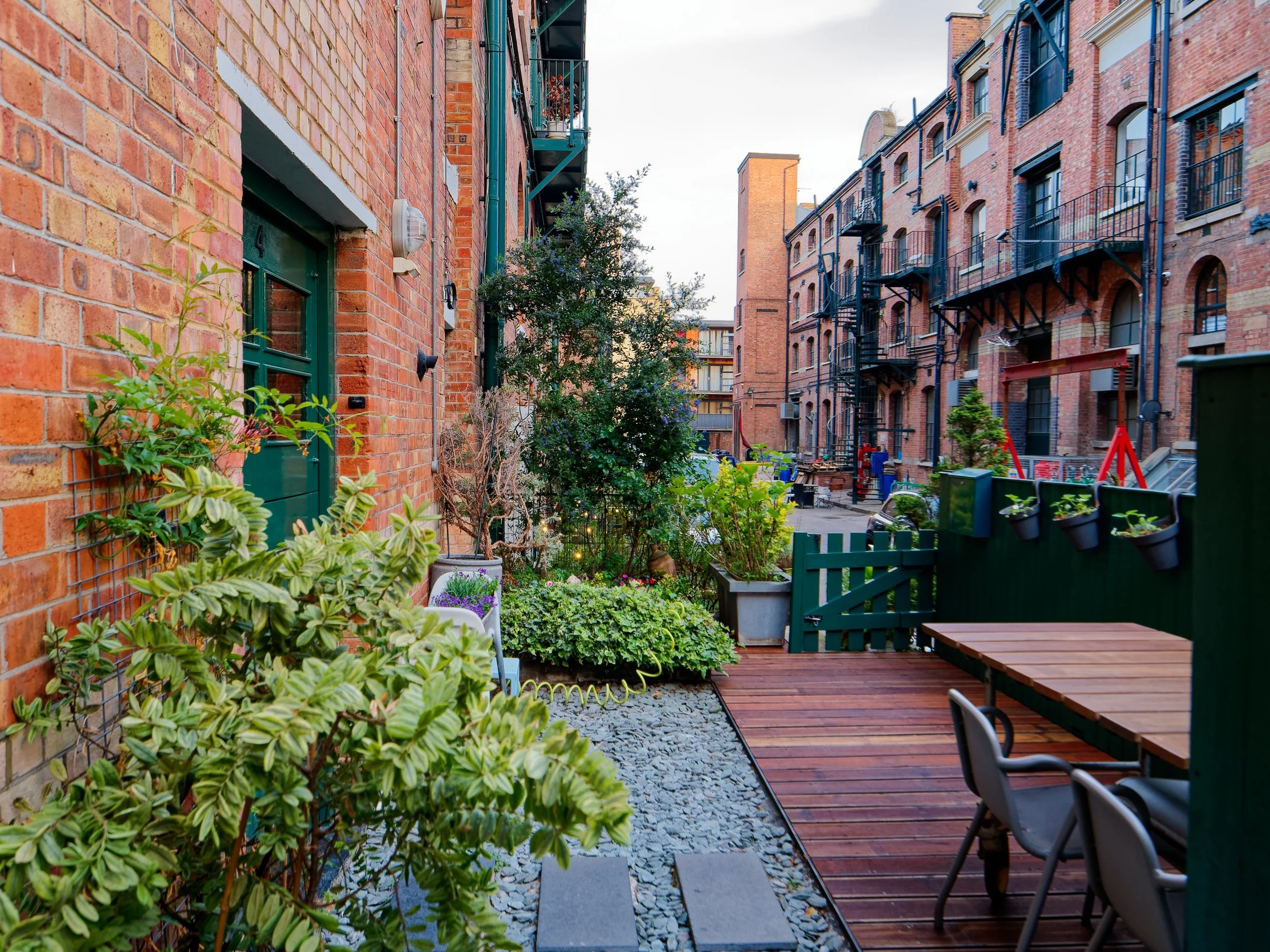 A cozy outdoor patio with wooden decking and a garden area filled with various plants and shrubs. Potted plants are on the green fence, and there are chairs and tables for seating. Surrounding brick buildings with fire escapes are visible in the back