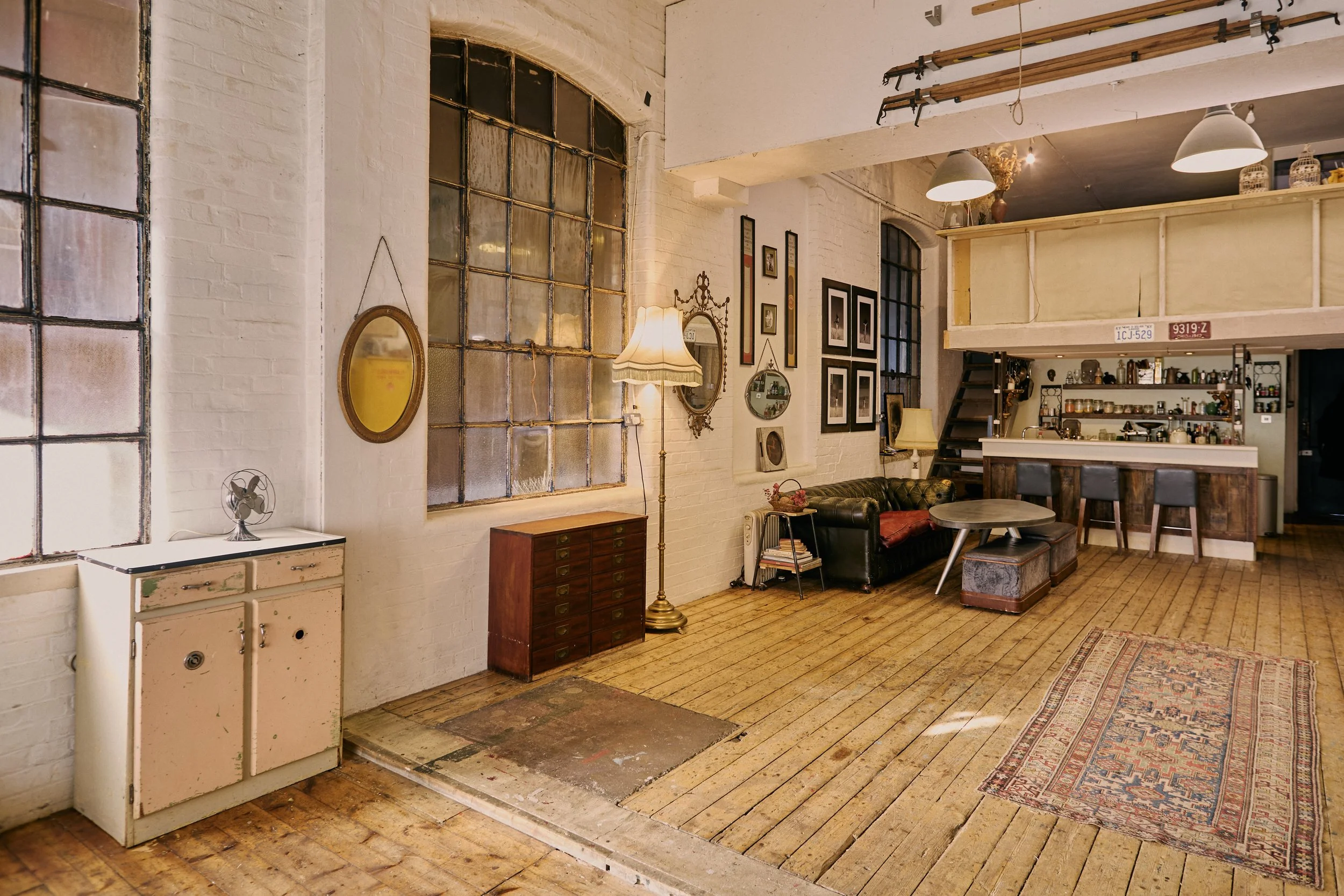 Interior of a spacious, rustic living room with wooden floors, vintage furniture, and eclectic decorations. Large industrial windows, a black leather sofa, framed pictures, a standing lamp with a shade, a small wooden cabinet, and a bar area with sto