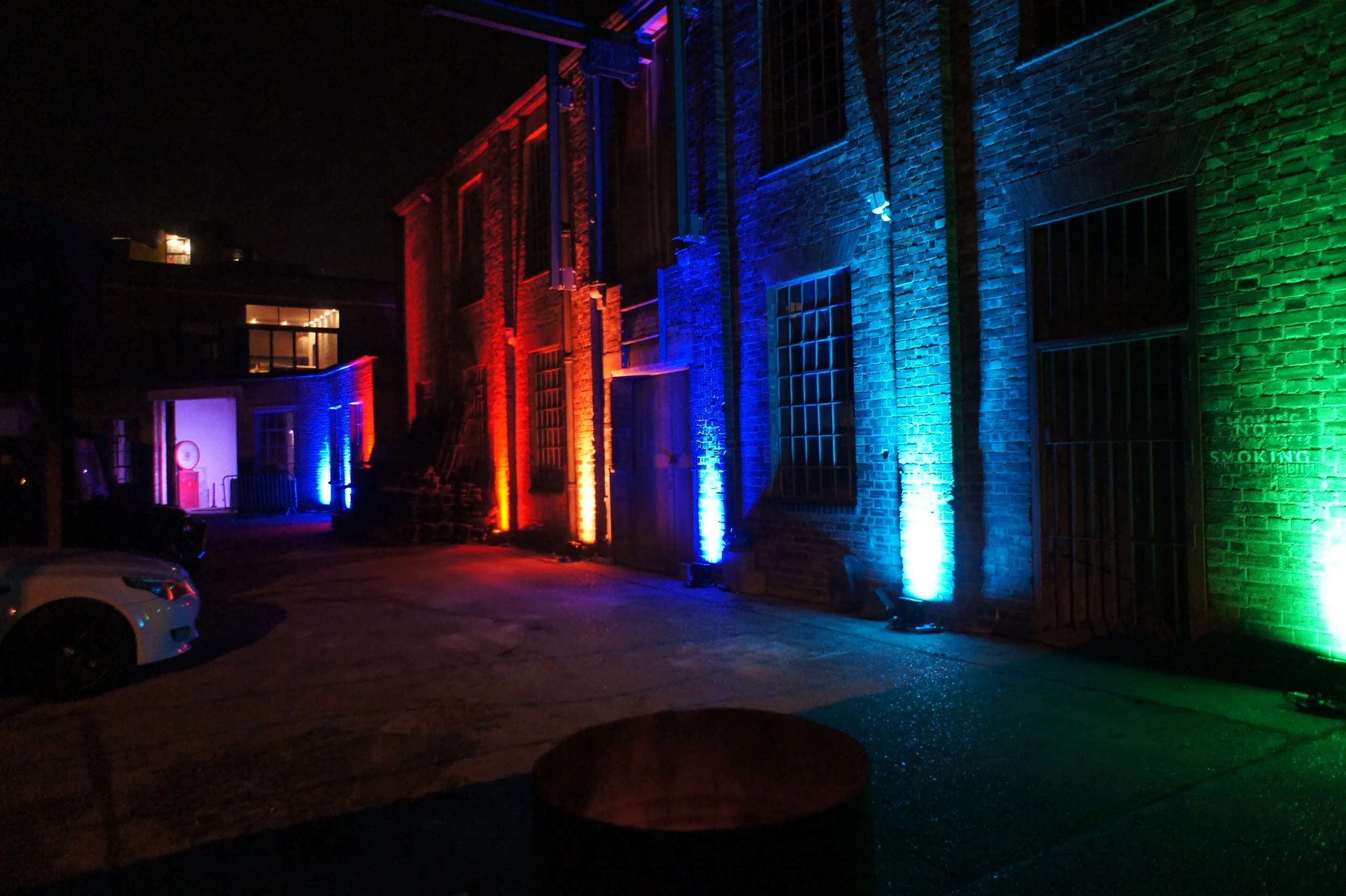 A nighttime scene of a brick building illuminated by colorful lights in red, blue, and green, with parking and a garage door visible.