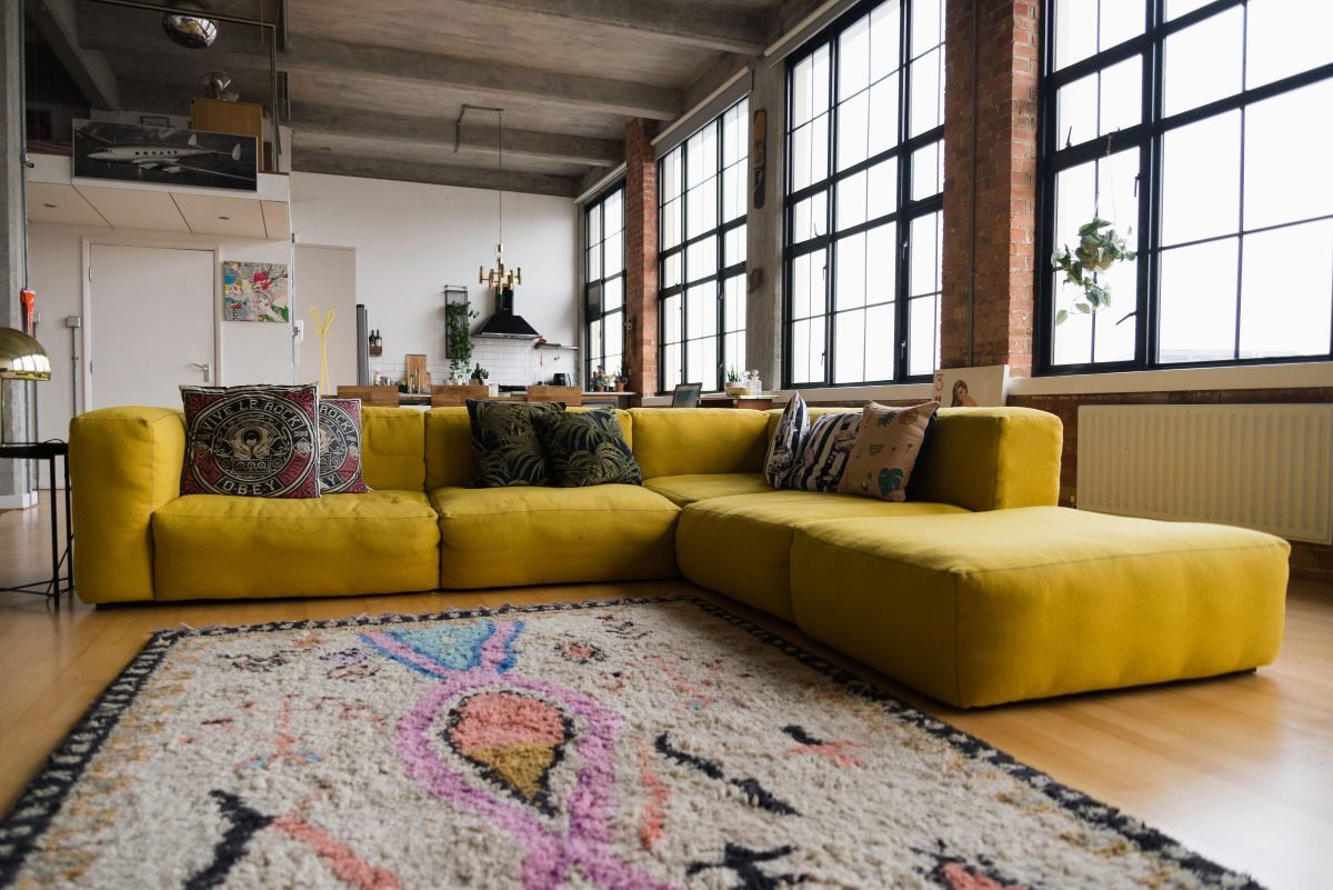 A modern living room with a large yellow sectional sofa, colorful throw pillows, a patterned area rug, and large windows with black frames and brick accents.