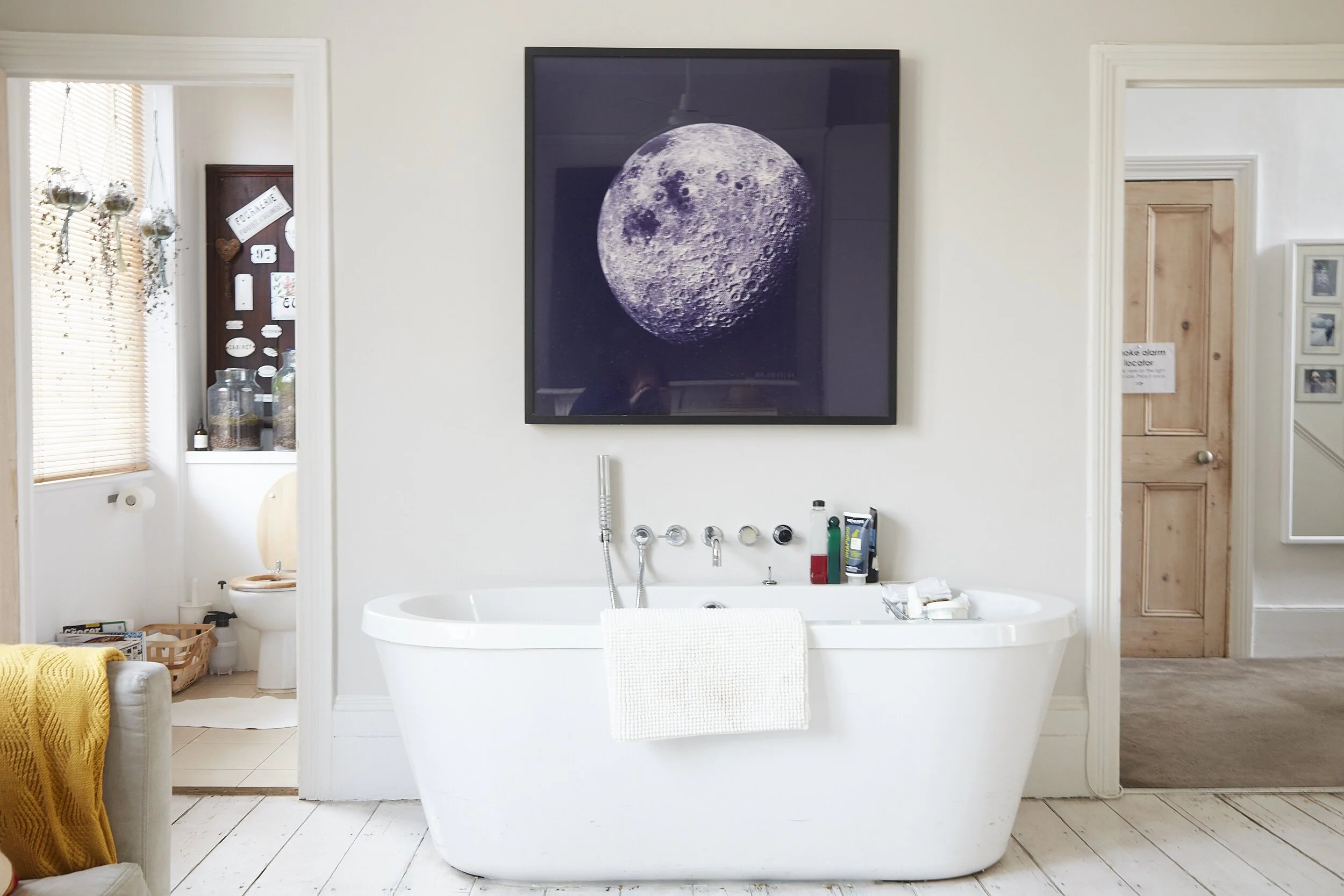 A bathroom with a white bathtub in front of a cream wall, a large framed moon photograph above the bathtub, and a small towel hanging on the edge. To the left, a doorway leads to a toilet in a small room with a window and blinds. To the right, anothe