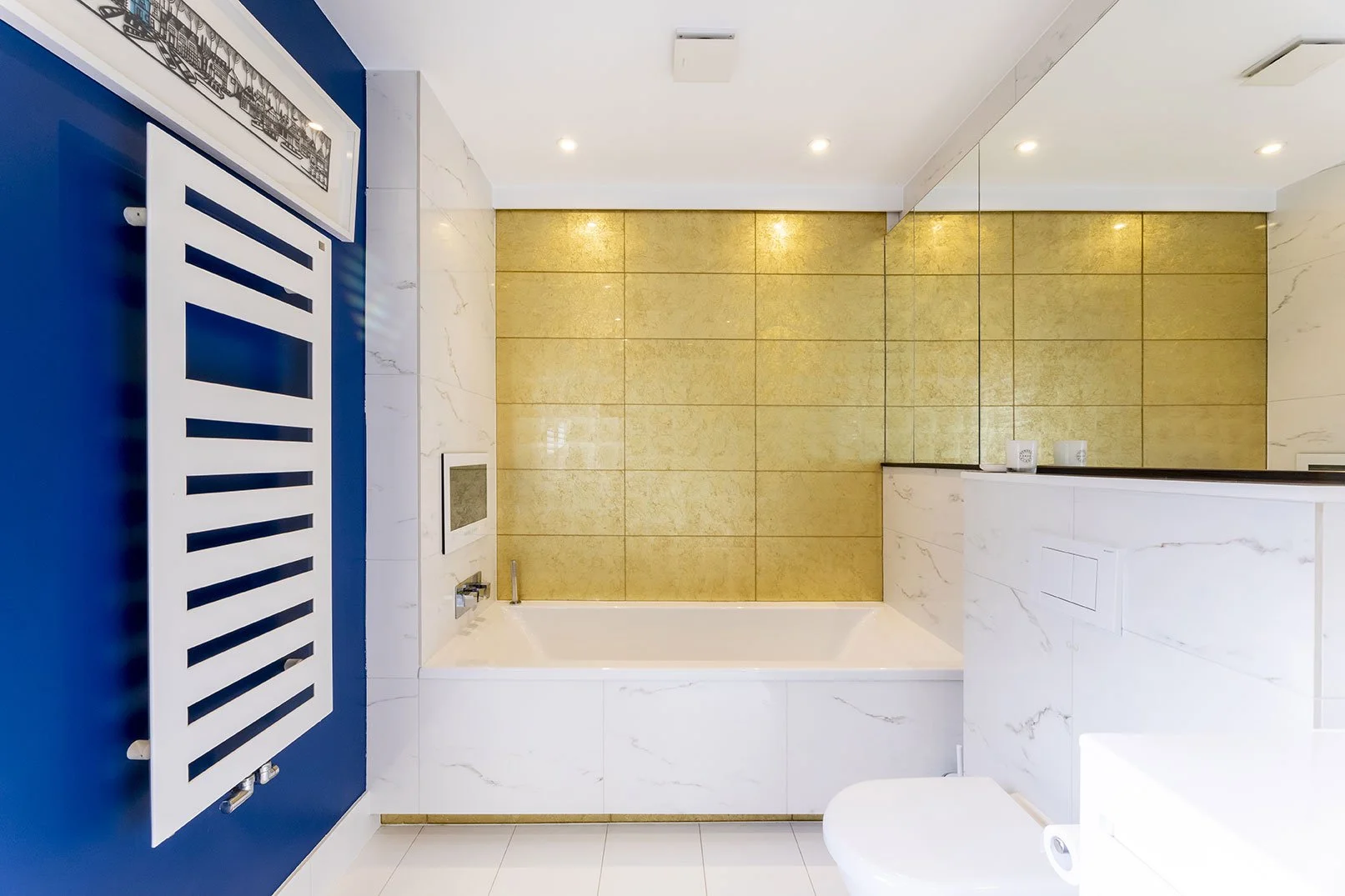 Modern bathroom with white marble walls, a blue accent wall with a radiator, a bathtub, a wall-mounted control panel, and a partial view of a toilet. Gold tiles are on the back wall and ceiling lights are installed for illumination.