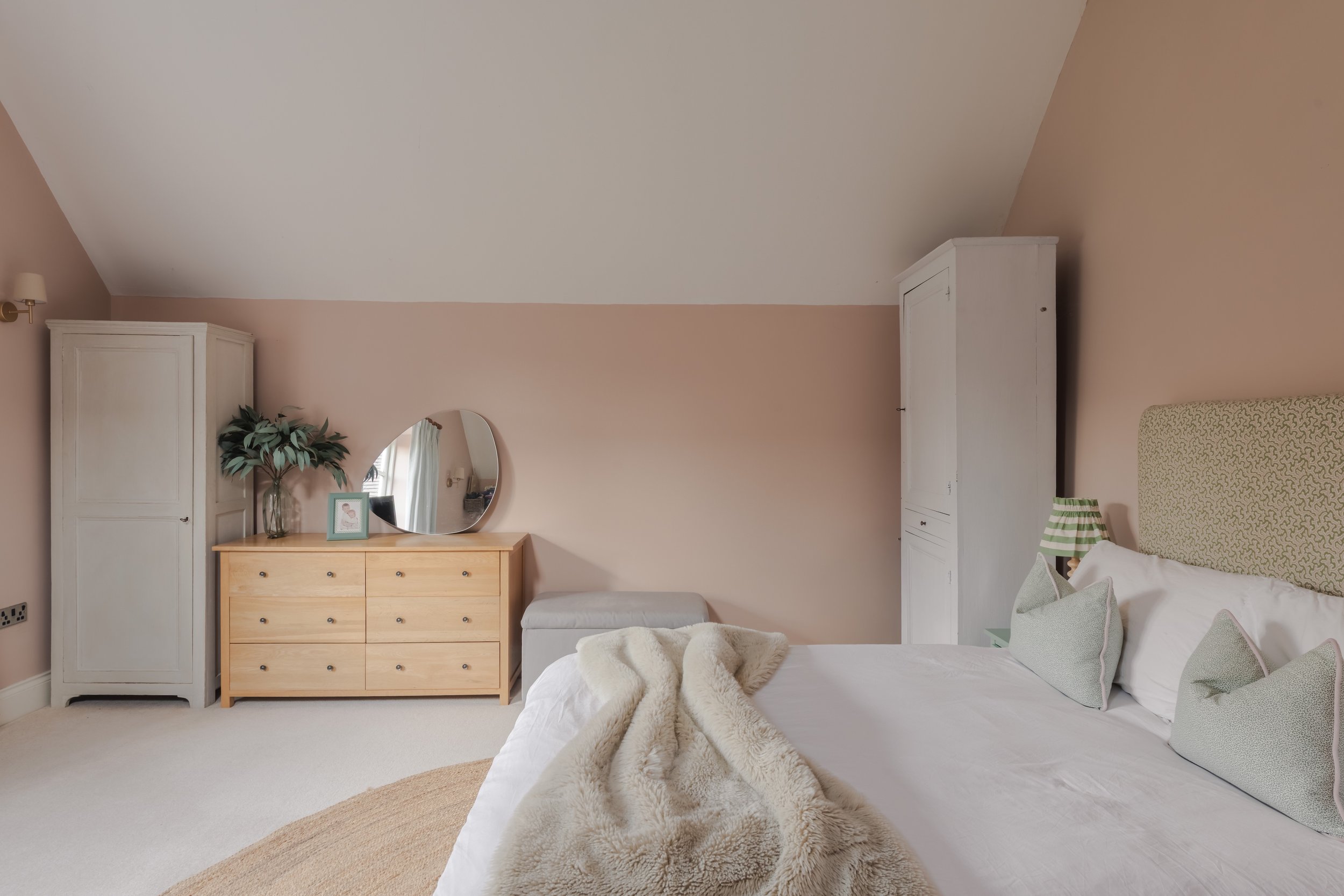 A cozy bedroom with a sloped ceiling, featuring a bed with light green pillows, a beige headboard, a nightstand with a striped lamp, a white wardrobe, a beige dresser with a mirror, a small framed picture, a vase with greenery, and a gray bench.