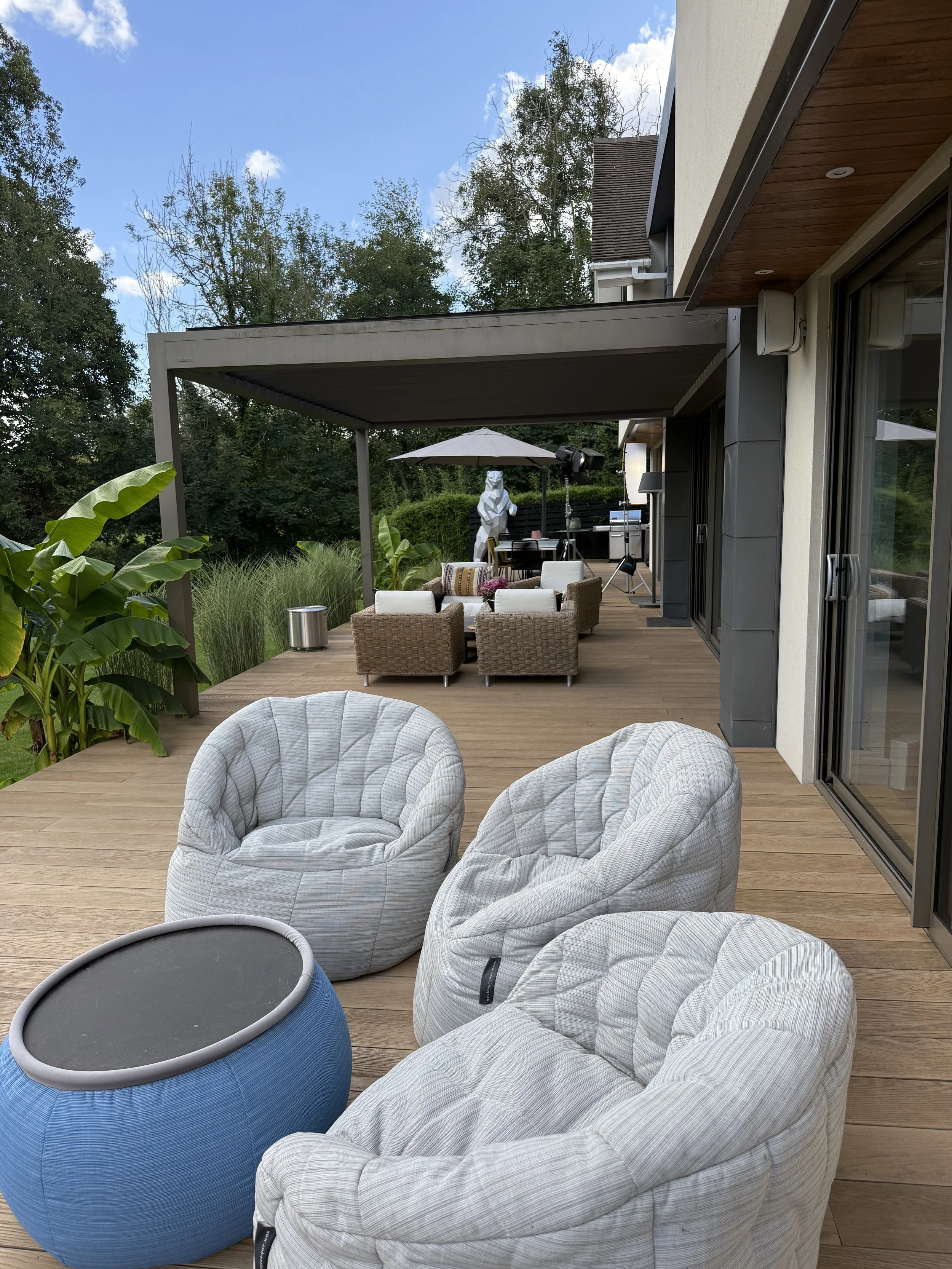 An outdoor patio area with cushioned chairs, a blue side table, and a seating area with wicker chairs, an umbrella, and a statue in the background, surrounded by greenery.