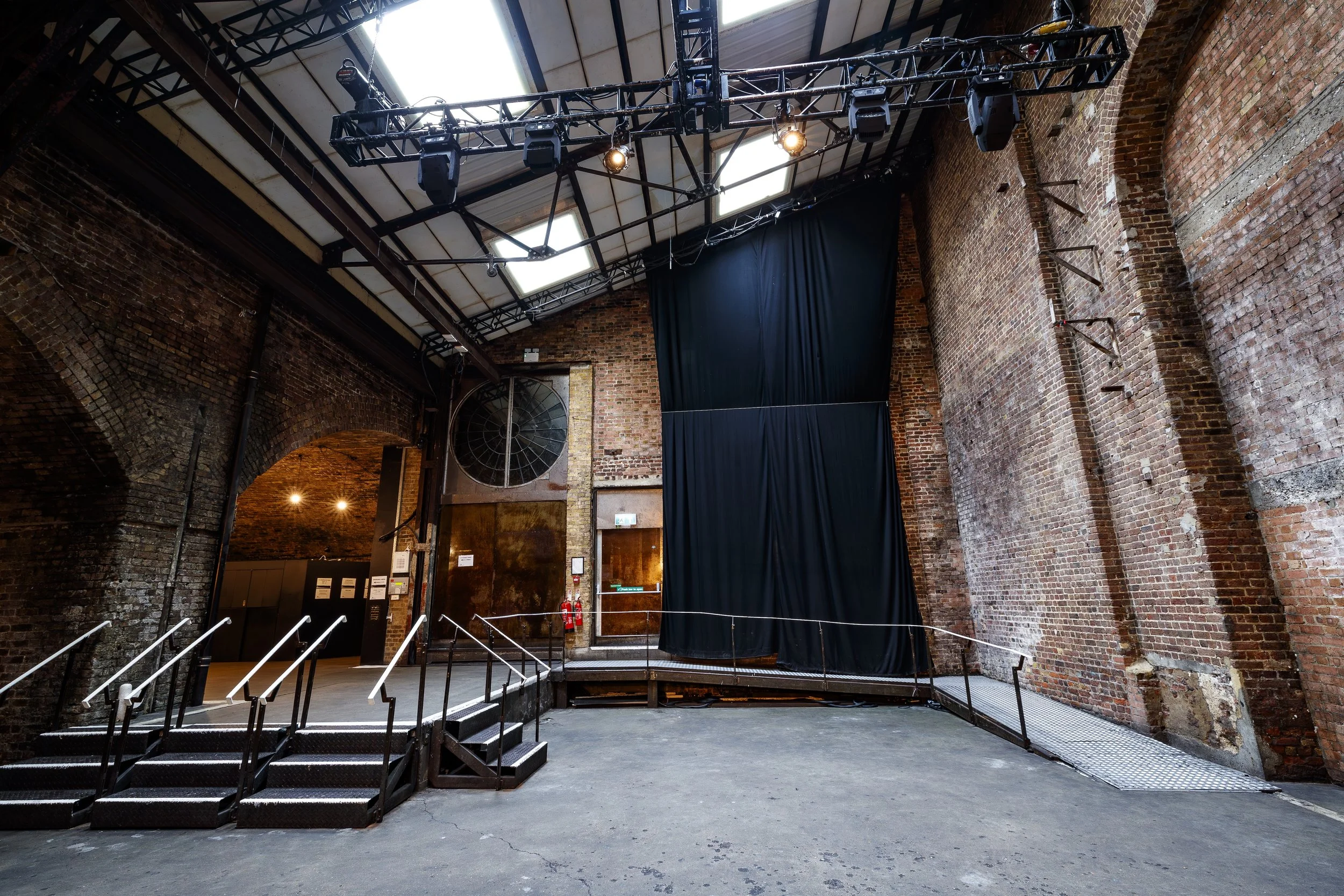 Interior of a converted industrial building with brick walls, a black stage curtain, metal stairs, and stage lighting equipment hanging from the ceiling.