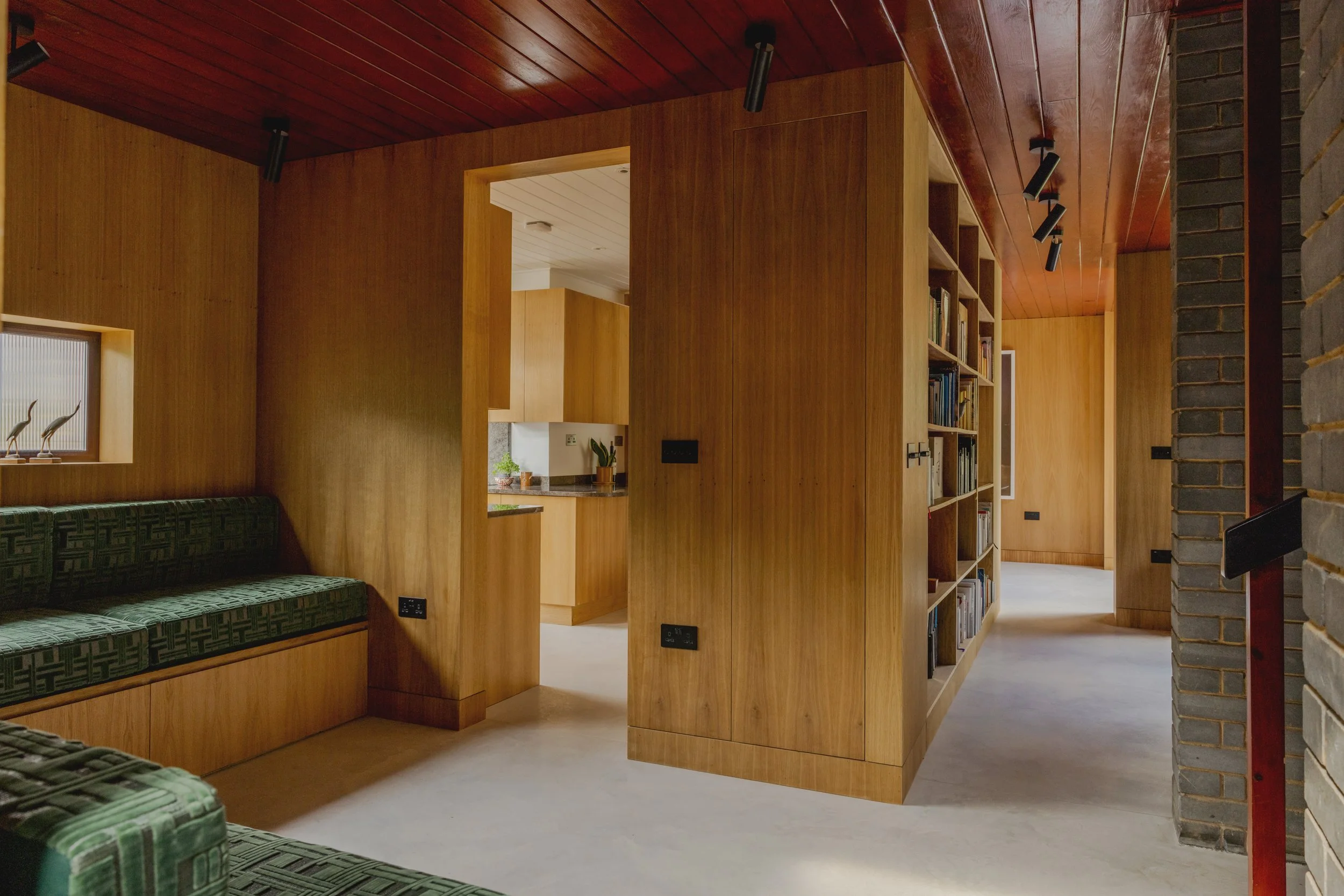 Interior view of a house showing wooden walls and ceiling, a bookshelf, and a seating area with green patterned furniture.