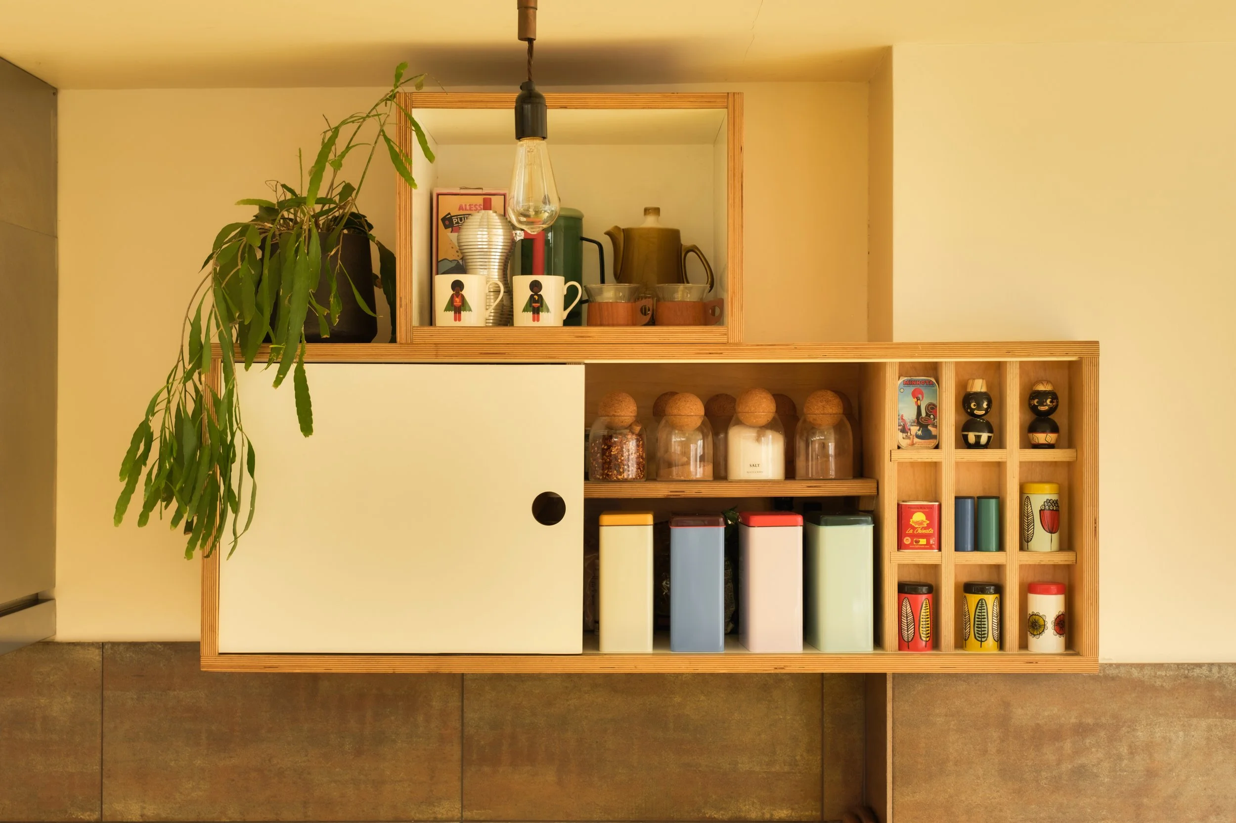 A wooden wall-mounted kitchen cabinet with a sliding door on the left side, containing various jars and containers, with a plant and decorative items on top, and shelves with colorful tins and small decorative figures.