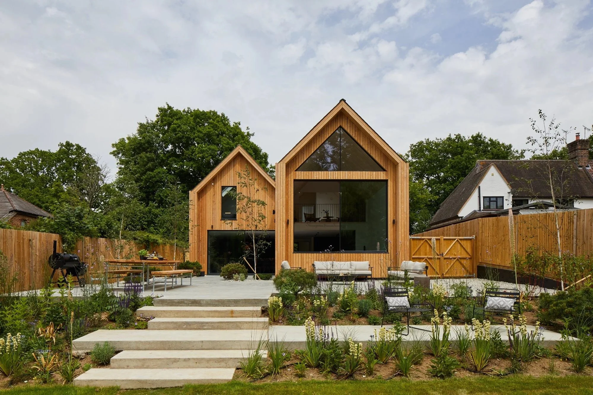 Modern house with wooden exterior, large glass windows, surrounding garden, outdoor seating, and wooden fence.